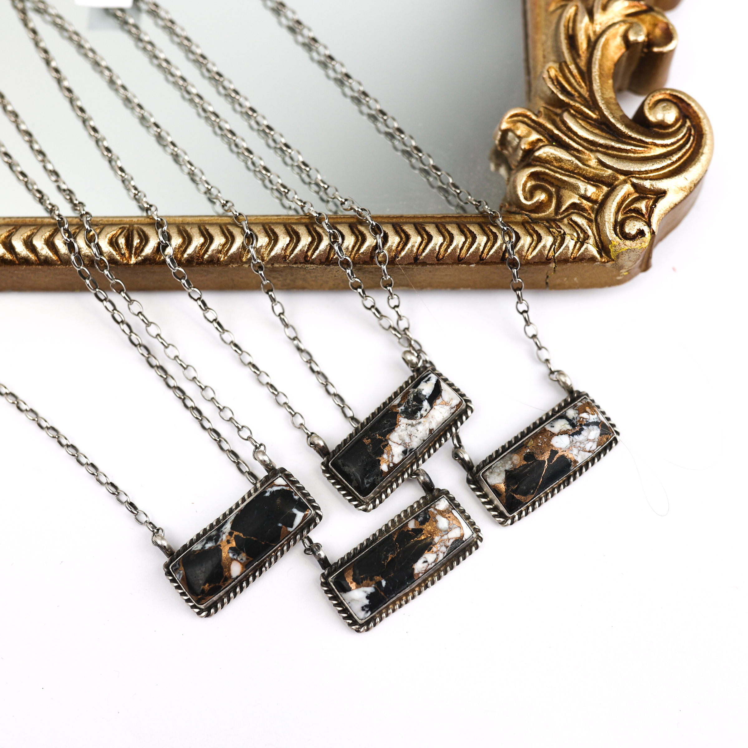 Four silver chain necklaces with rectangle bars that have a white buffalo stone. These necklaces are pictured on a white background and partially laying on a mirror with a gold outline. 
