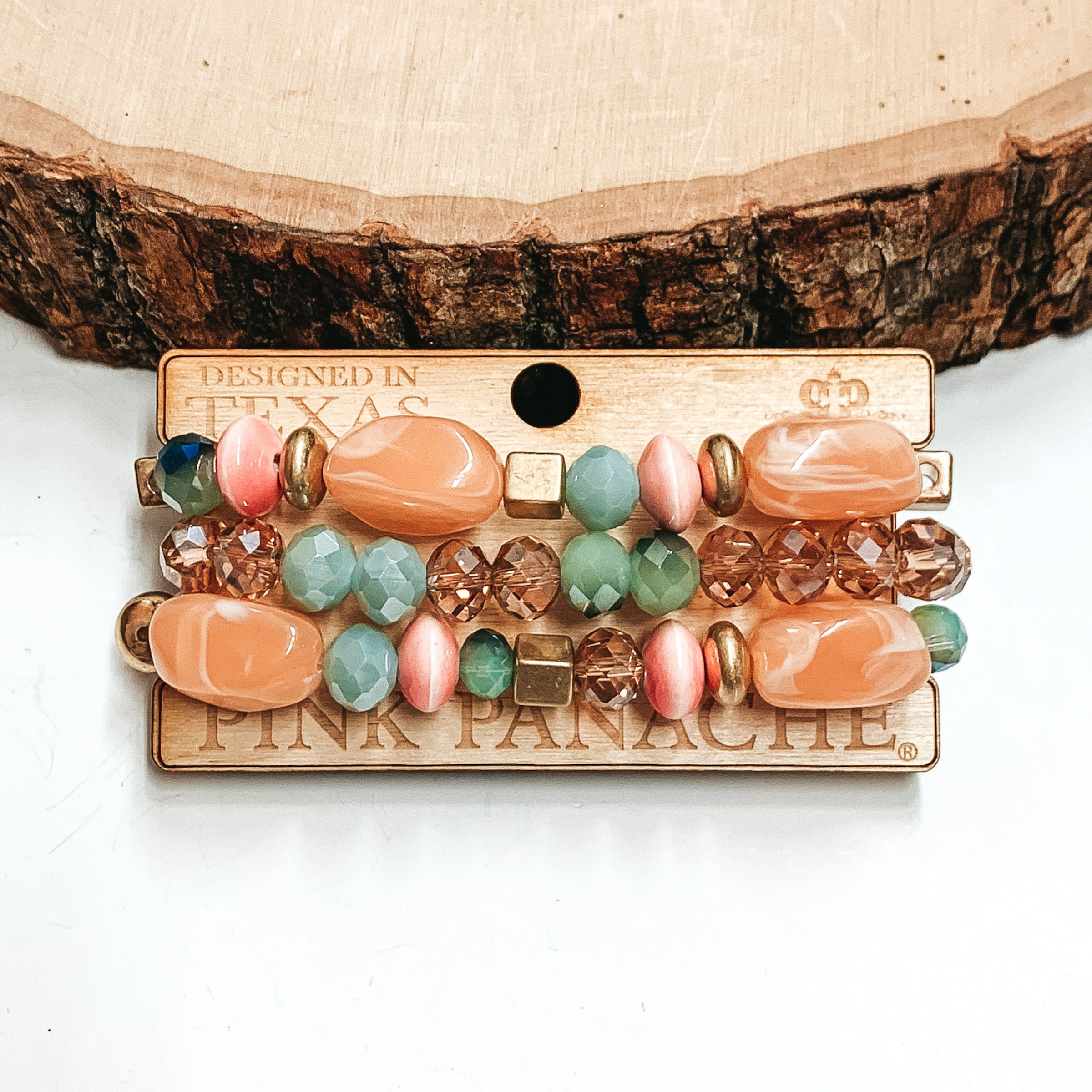 Peach, green, gold, and tan beaded bracelets. There are three bracelets pictured on a wood holder. These bracelets are pictured on front of a wood block on a white background. 