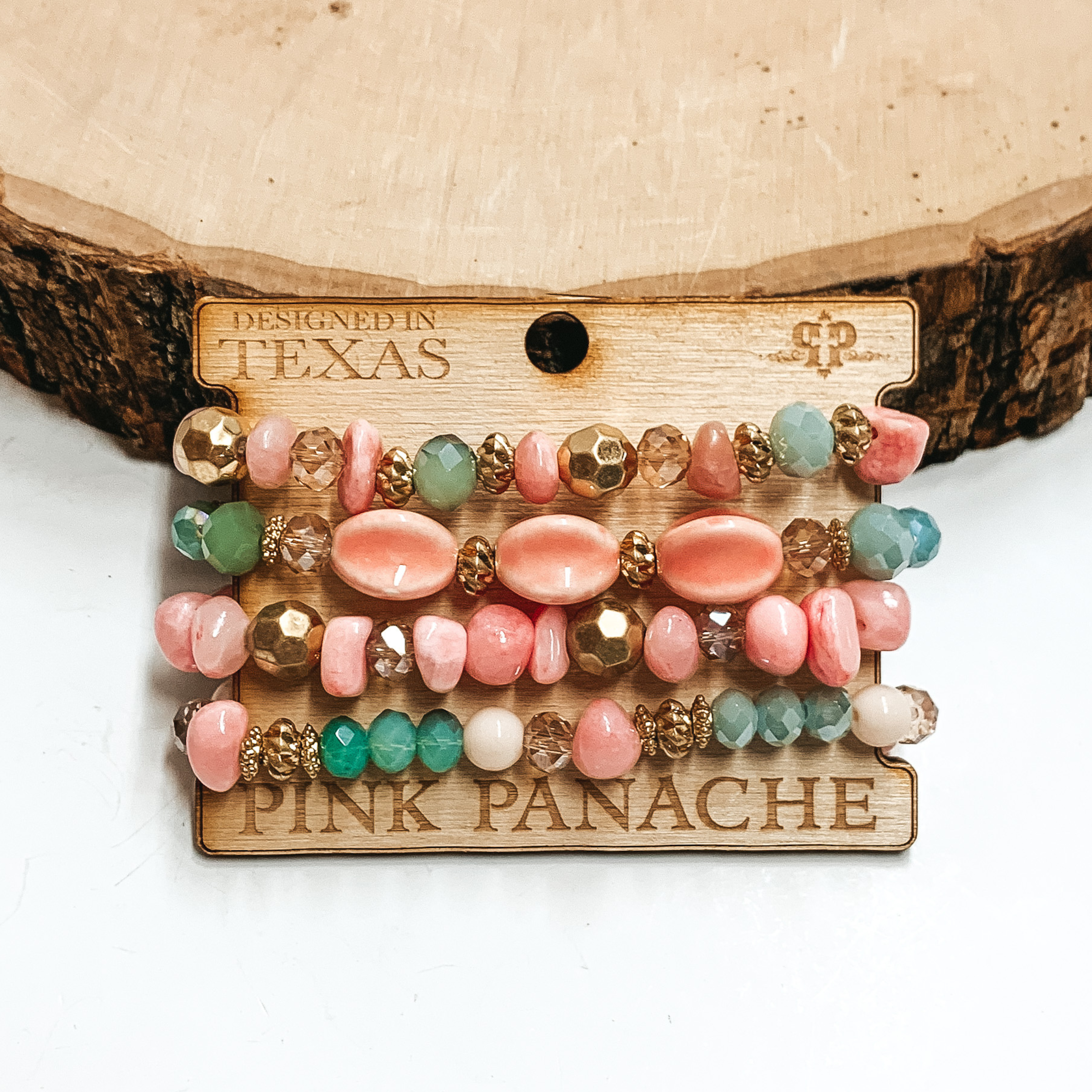 Peach, green, gold, and tan beaded bracelets. There are four bracelets pictured on a wood holder. These bracelets are pictured on front of a wood block on a white background. 