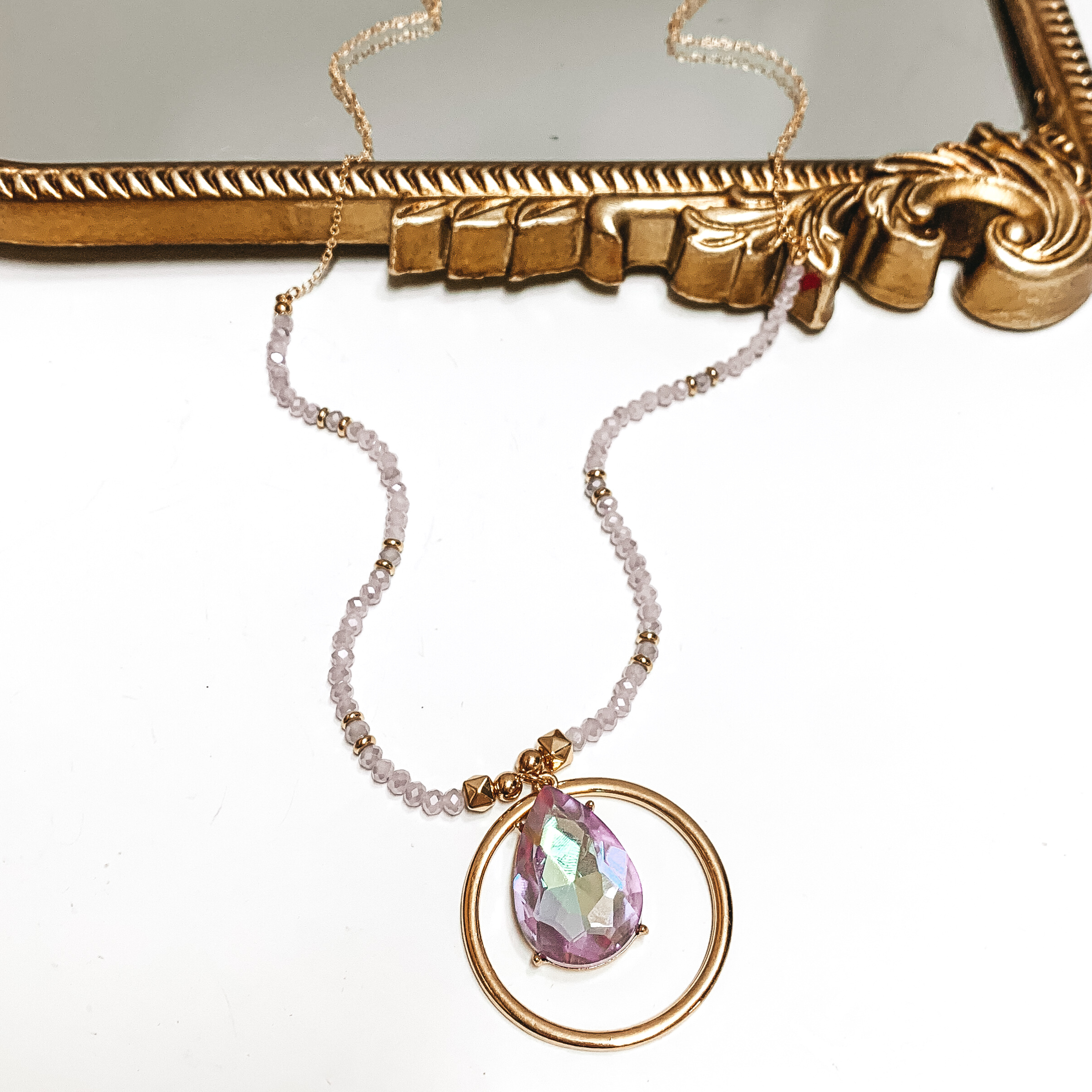 Gold and lavender beaded necklace with a gold, open circle pendant and hanging purple ab teardrop crystal. This necklace is pictured partially laying on a mirror on a white background. 
