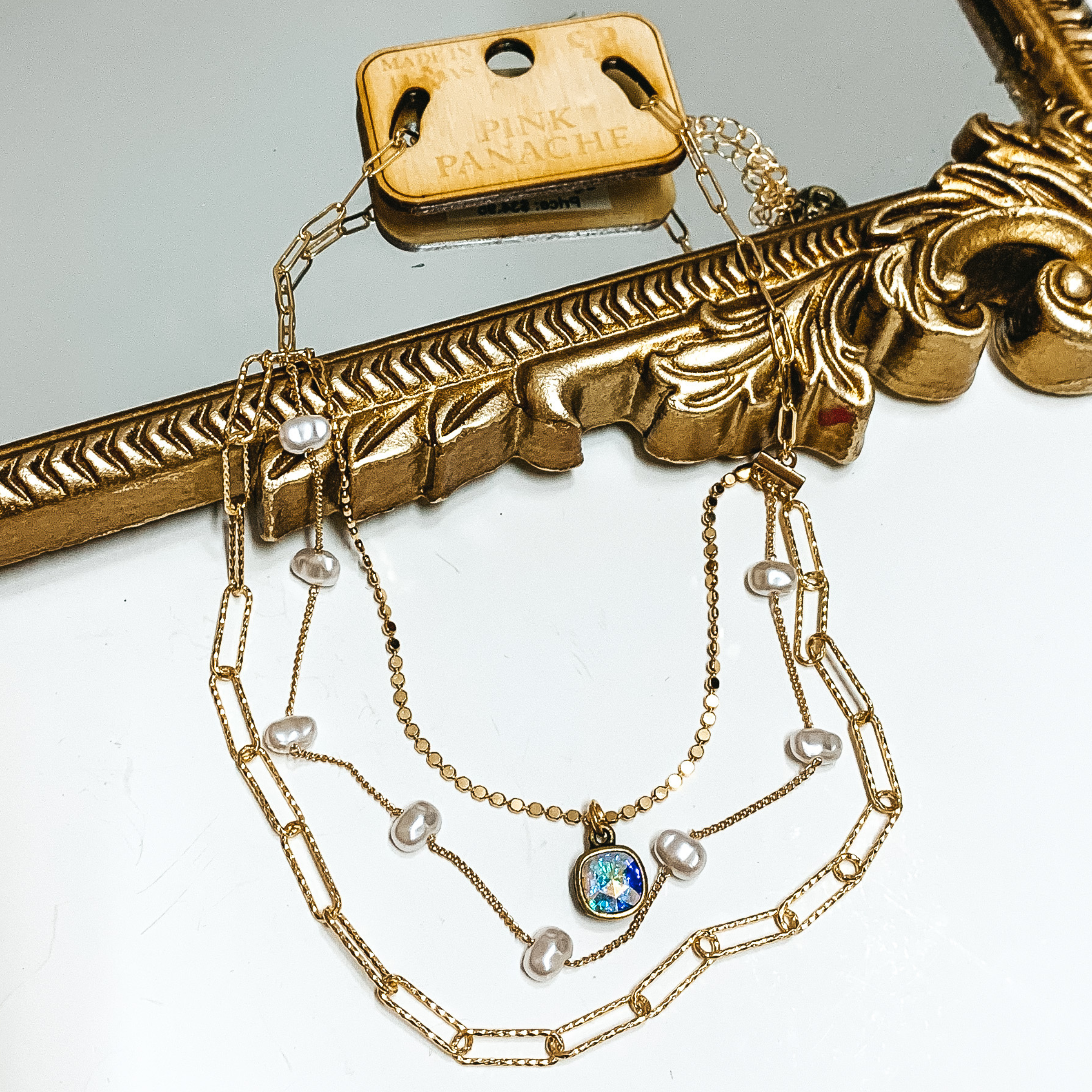 Three strand gold chain necklace. This necklace has white pearl spacers and a hanging ab cushion cut crystal. This necklace is pictured partially laying on a mirror on a white background. 