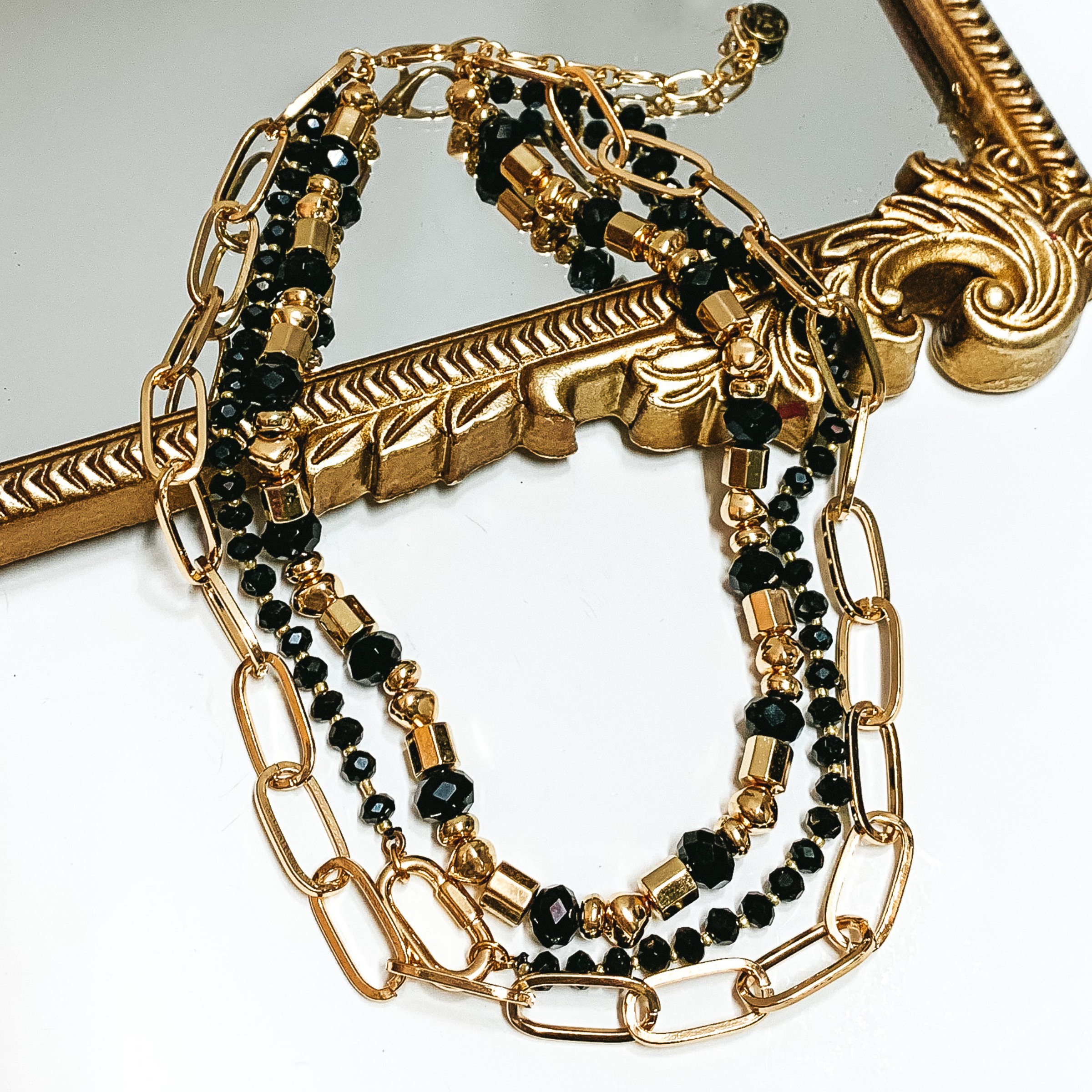 Black and gold beaded three strand necklace. This necklace includes a large, gold chain link strand. This necklace is pictured partially laying on a mirror on a white background. 