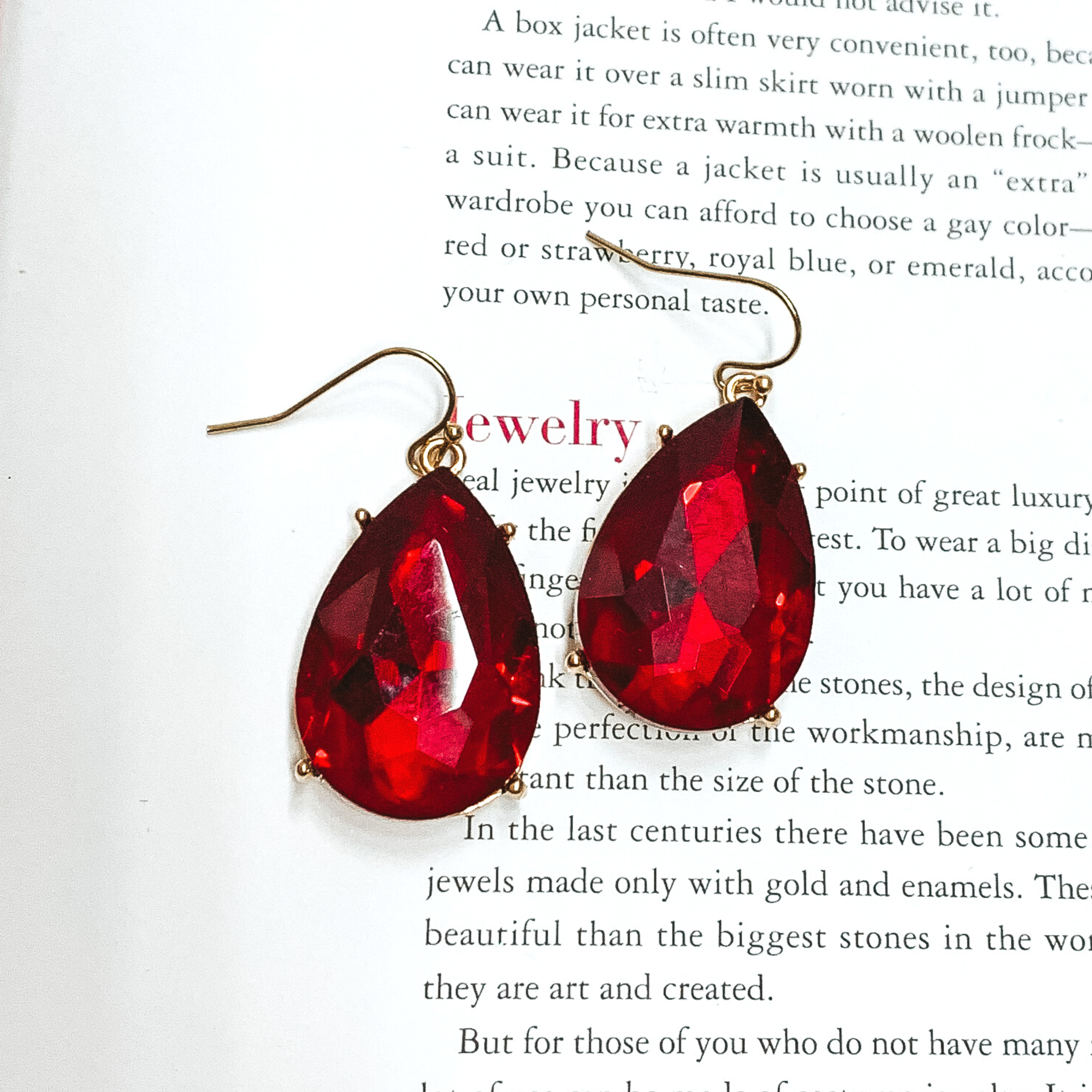 Gold fish hook earrings with a hanging red teardrop crystal. These earrings are pictured on an open book.