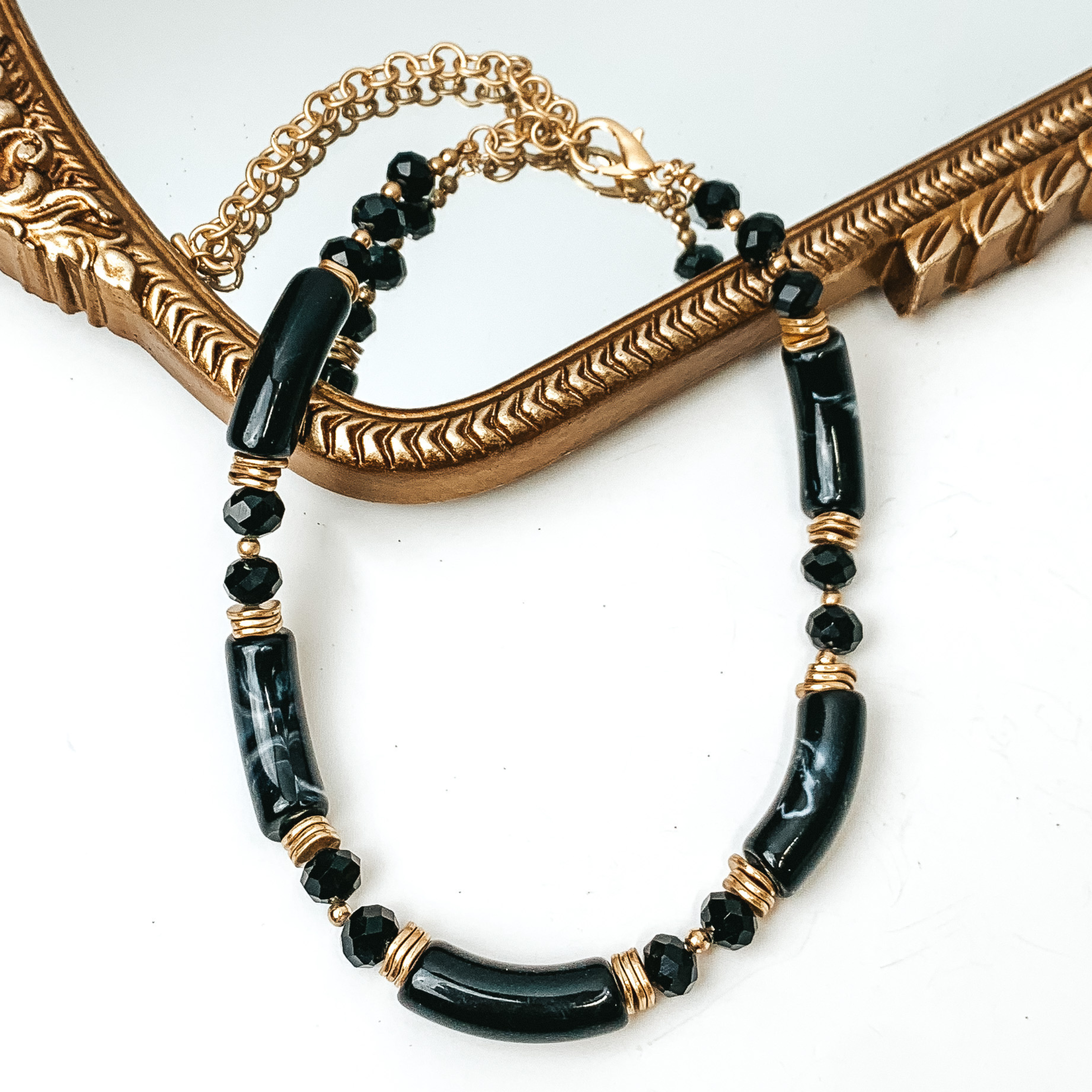 Black marble tube necklae with black crystal beads and gold bead spacers. This necklace is pictured partially laying on a gold mirror on a white background. 