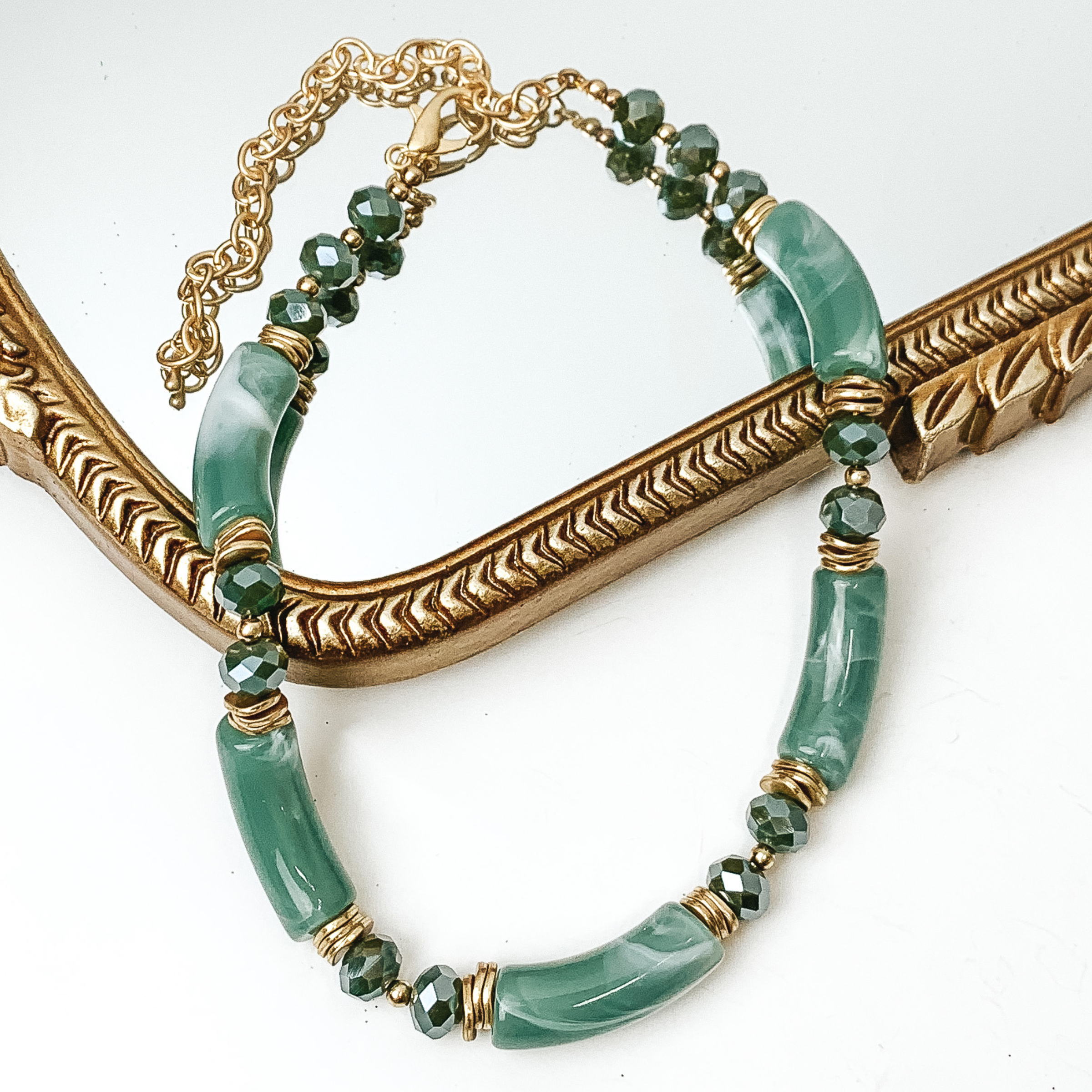 Green marble tube necklae with green crystal beads and gold bead spacers. This necklace is pictured partially laying on a gold mirror on a white background. 