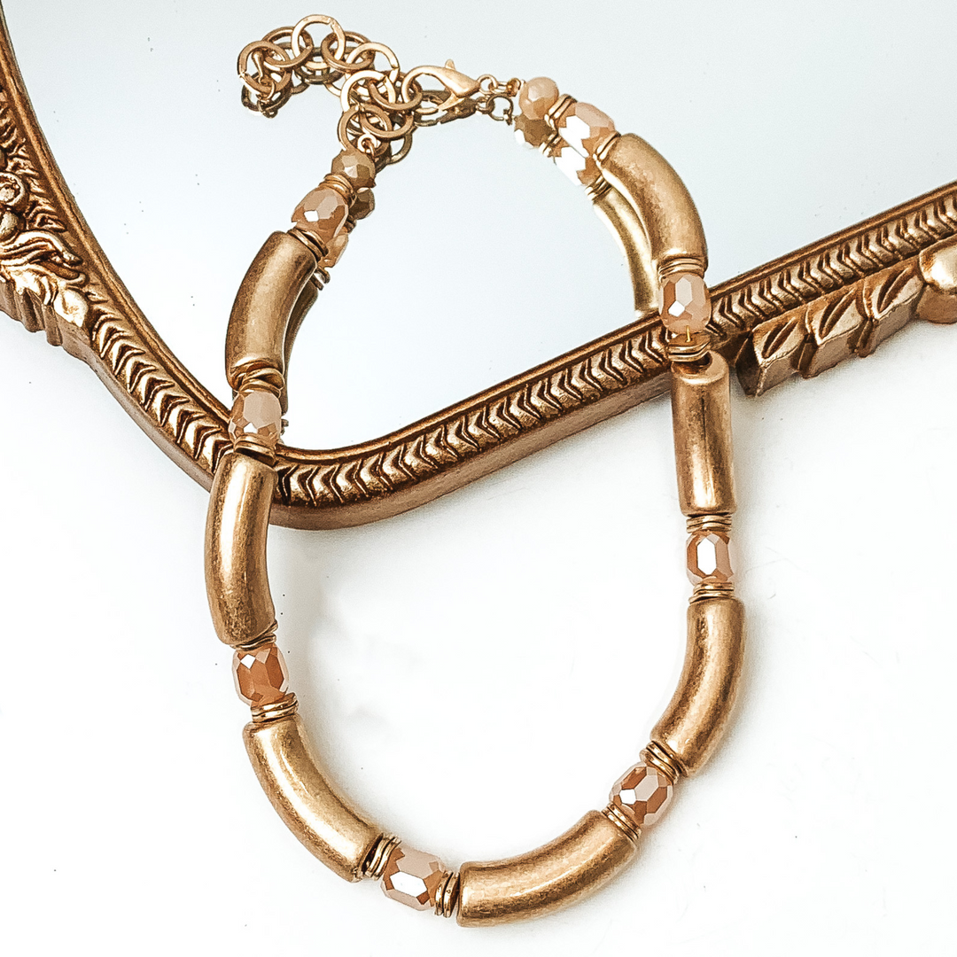 Gold tube necklae with beige crystal bead spacers. This necklace is pictured partially laying on a gold mirror on a white background. 