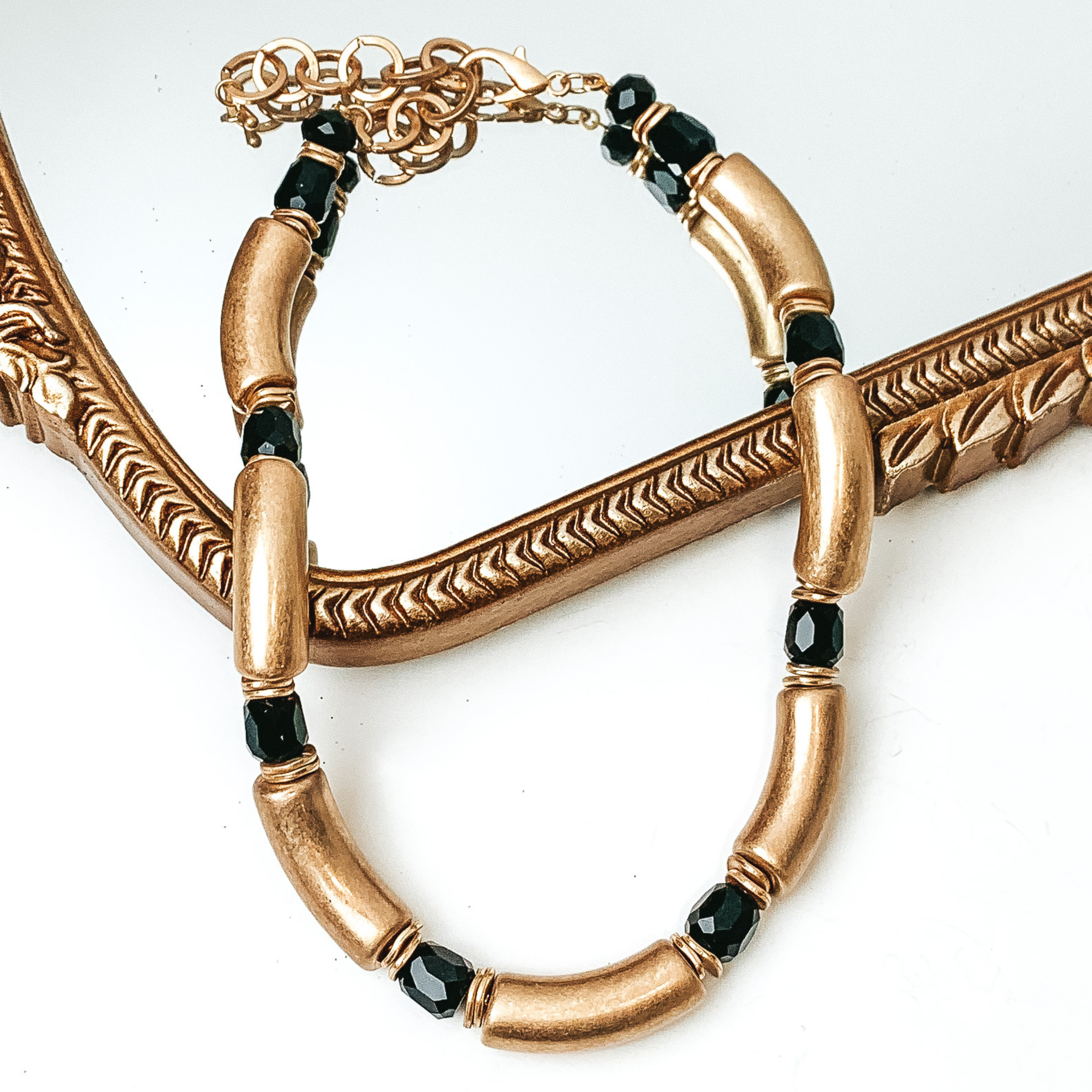 Gold tube necklae with black crystal bead spacers. This necklace is pictured partially laying on a gold mirror on a white background. 
