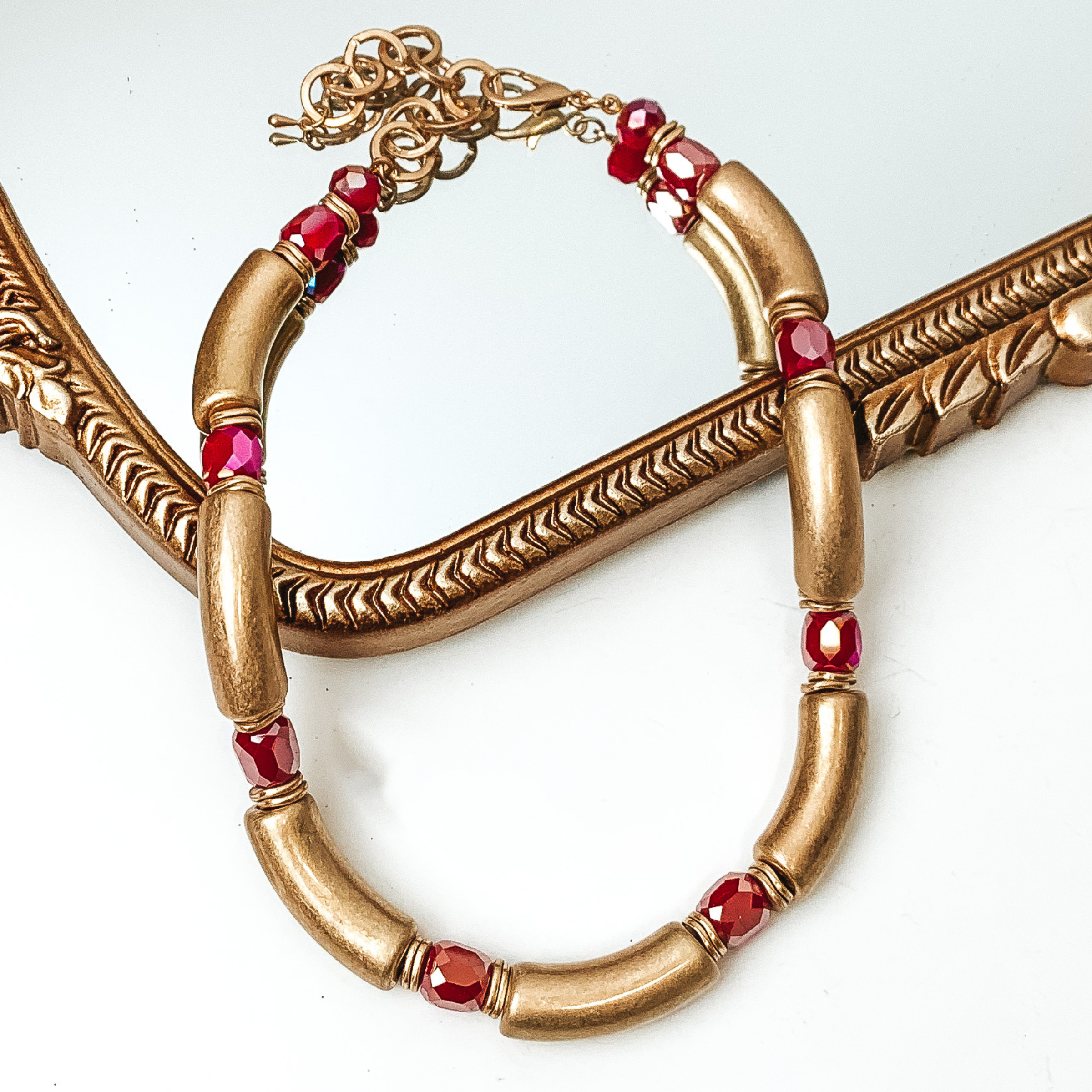 Gold tube necklae with red crystal bead spacers. This necklace is pictured partially laying on a gold mirror on a white background. 