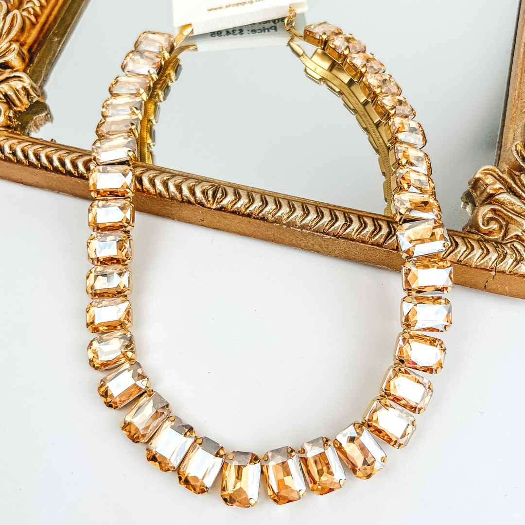 Beige, rectangle crystal necklace. This necklace is pictured laying on a gold mirror on a white background. 