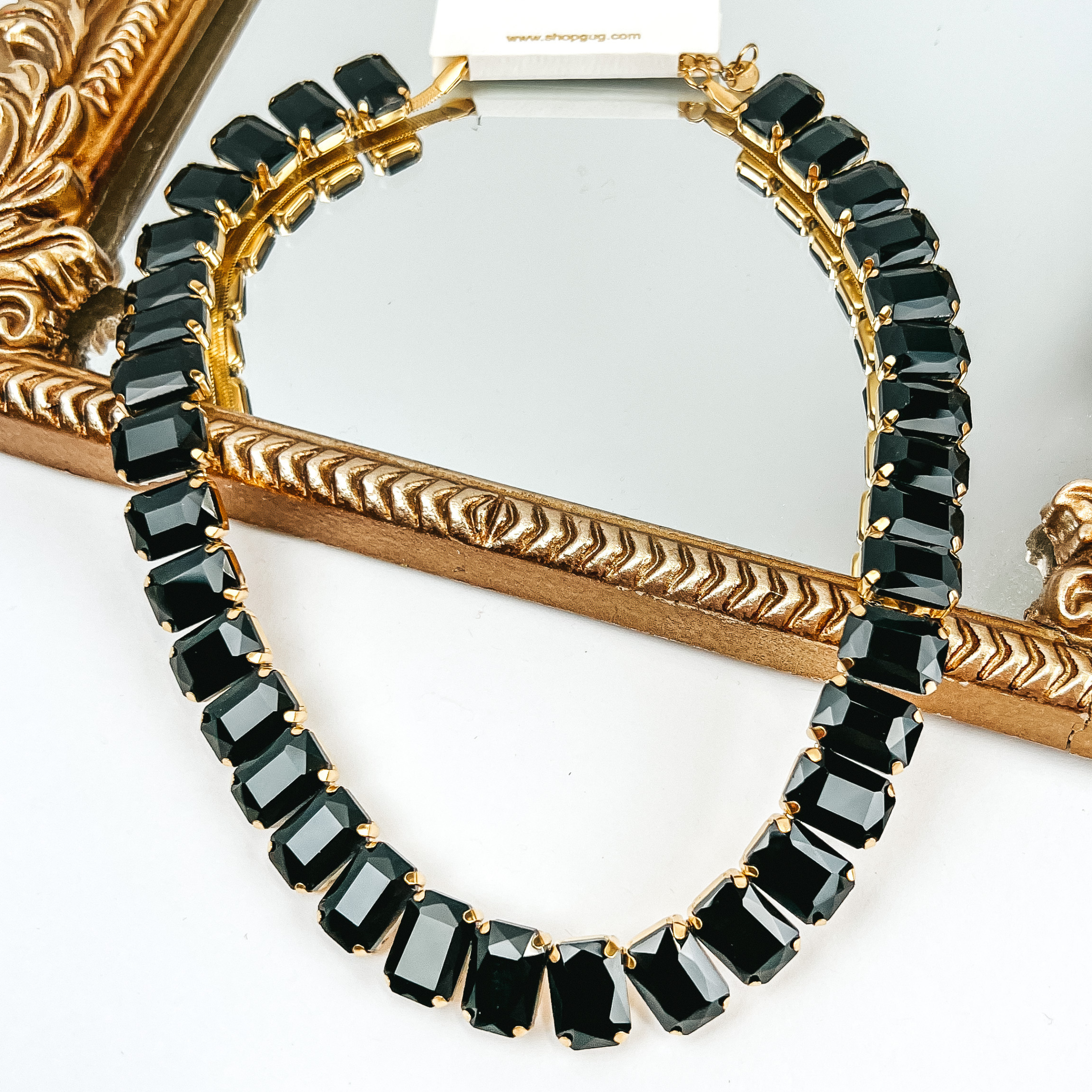 Black, rectangle crystal necklace. This necklace is pictured partially laying on a gold mirror on a white background. 