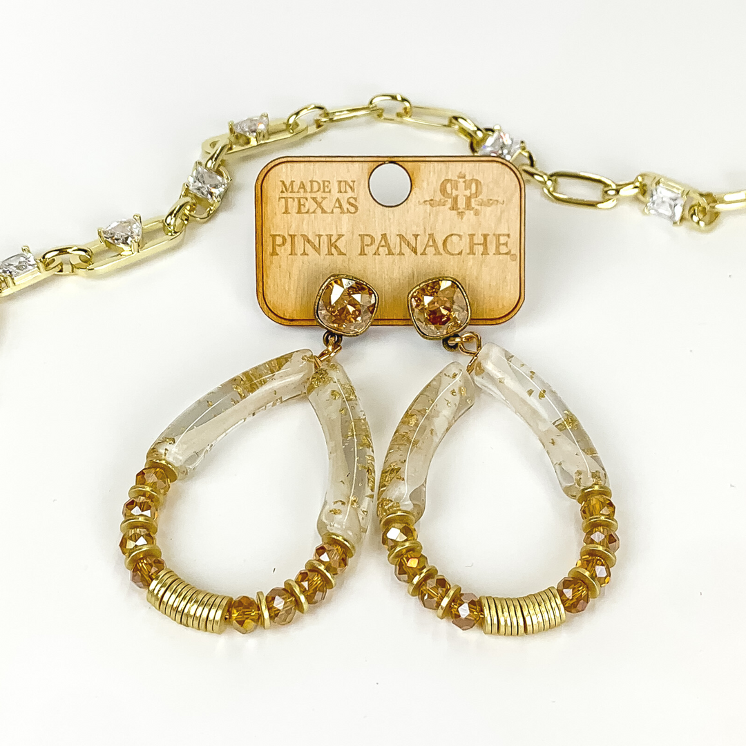 Tan square stud earrings with a gold and tan beaded teardrop pendant. The pendant includes gold disk beads, clear and gold flake tube beads, and tan crystal beads. These earrings are pictured on a white background with a gold chain behind the earrings. 