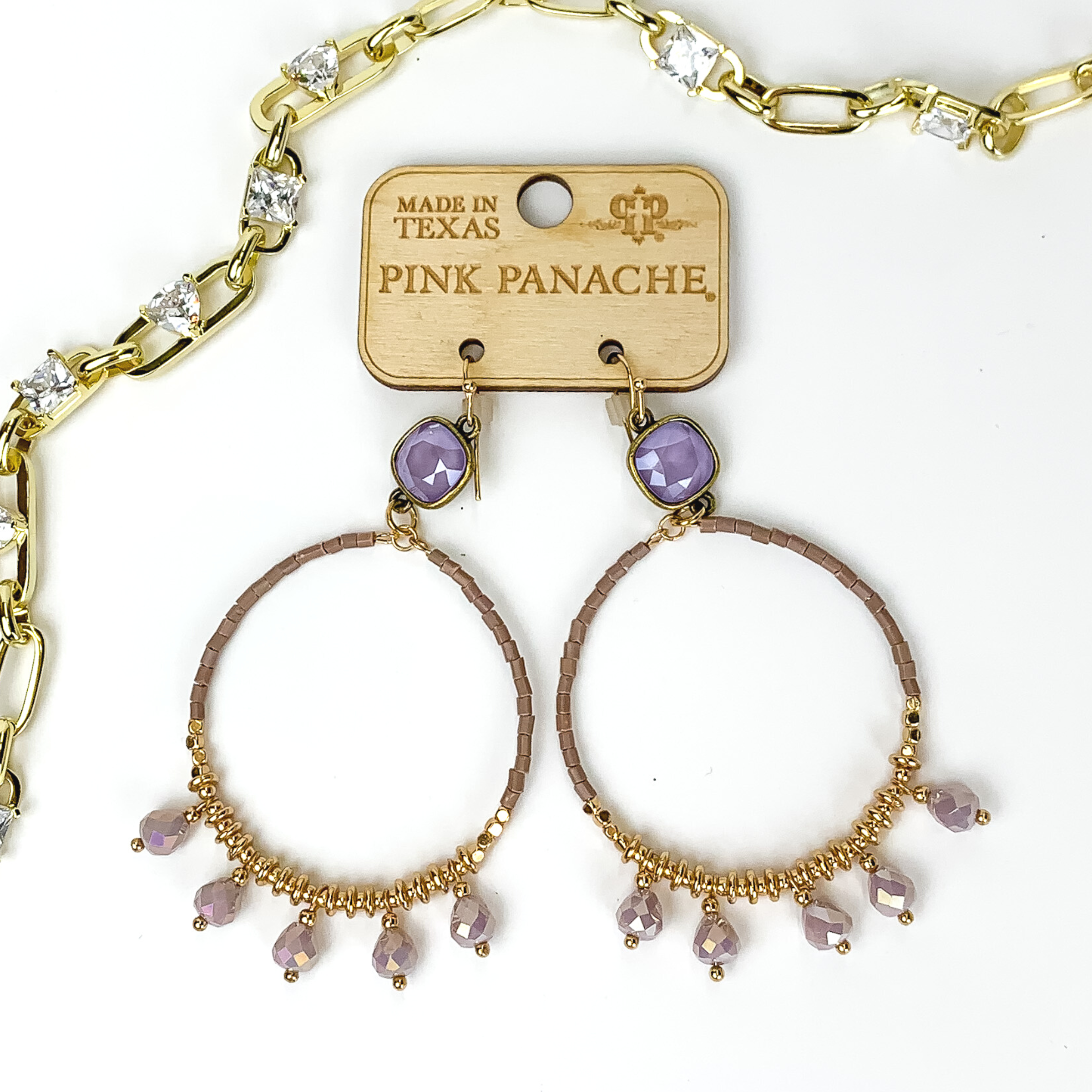 Lavender crystal square stud drop earrings with lavender beaded circle pendant. The circle pendant includes lavender beads, gold beads, and lilac hanging charms. These earrings are pictured on a top of a mauve colored book on a white background. 