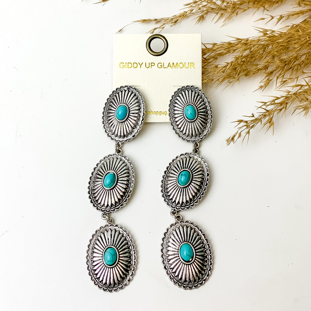 Three tiered silver, oval concho earrings. The conchos also include a center turquoise stone. These earrings are pictured on a white background with tan pompous grass in the top right corner.