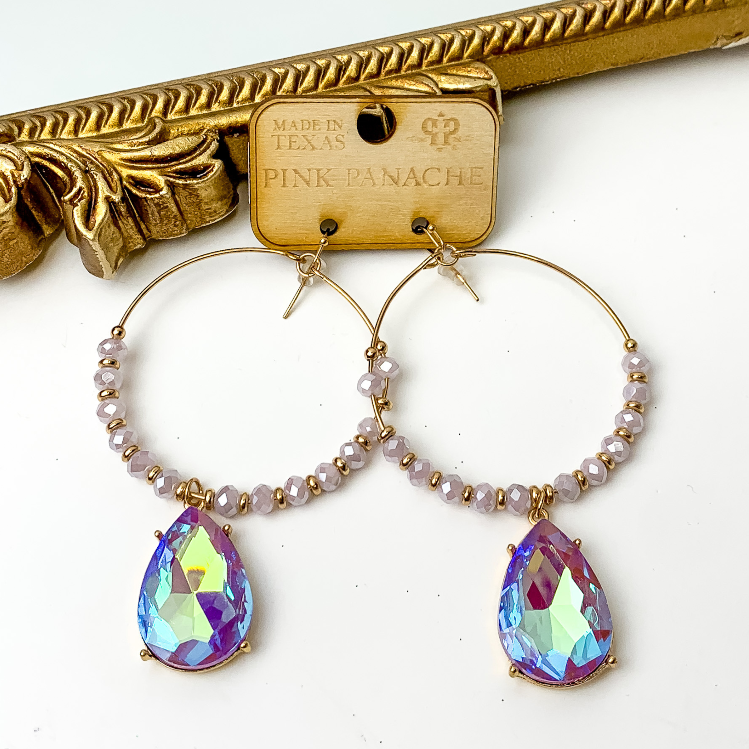 Large gold hoop earrings with lavender and gold beads. These hoops also include a teardrop purple ab crystal charm. These earrings are pictured on a white background with a gold mirror in the top left corner. 