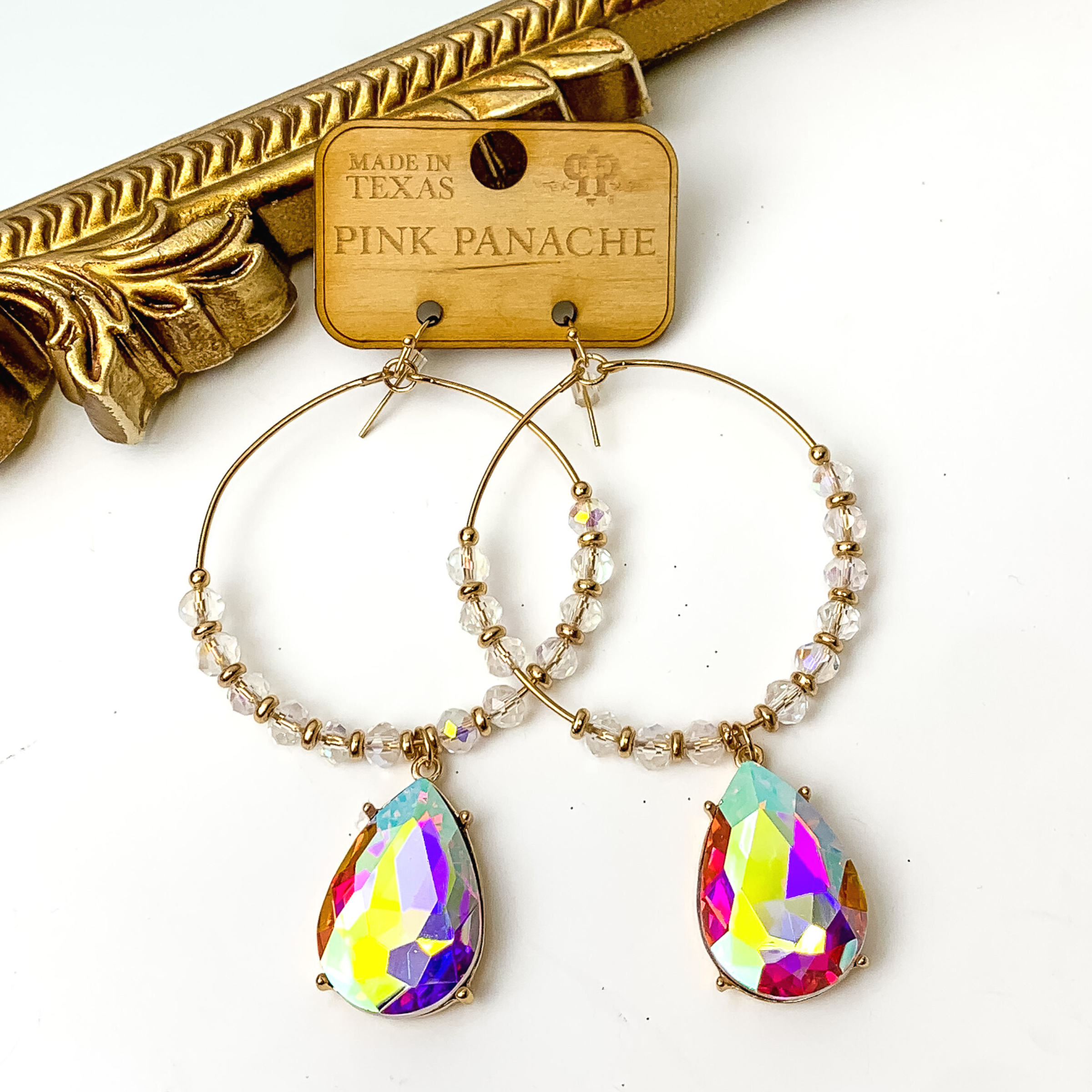 Large gold hoop earrings with clear ab and gold beads. These hoops also include a teardrop ab crystal charm. These earrings are pictured on a white background with a gold mirror in the top left corner. 