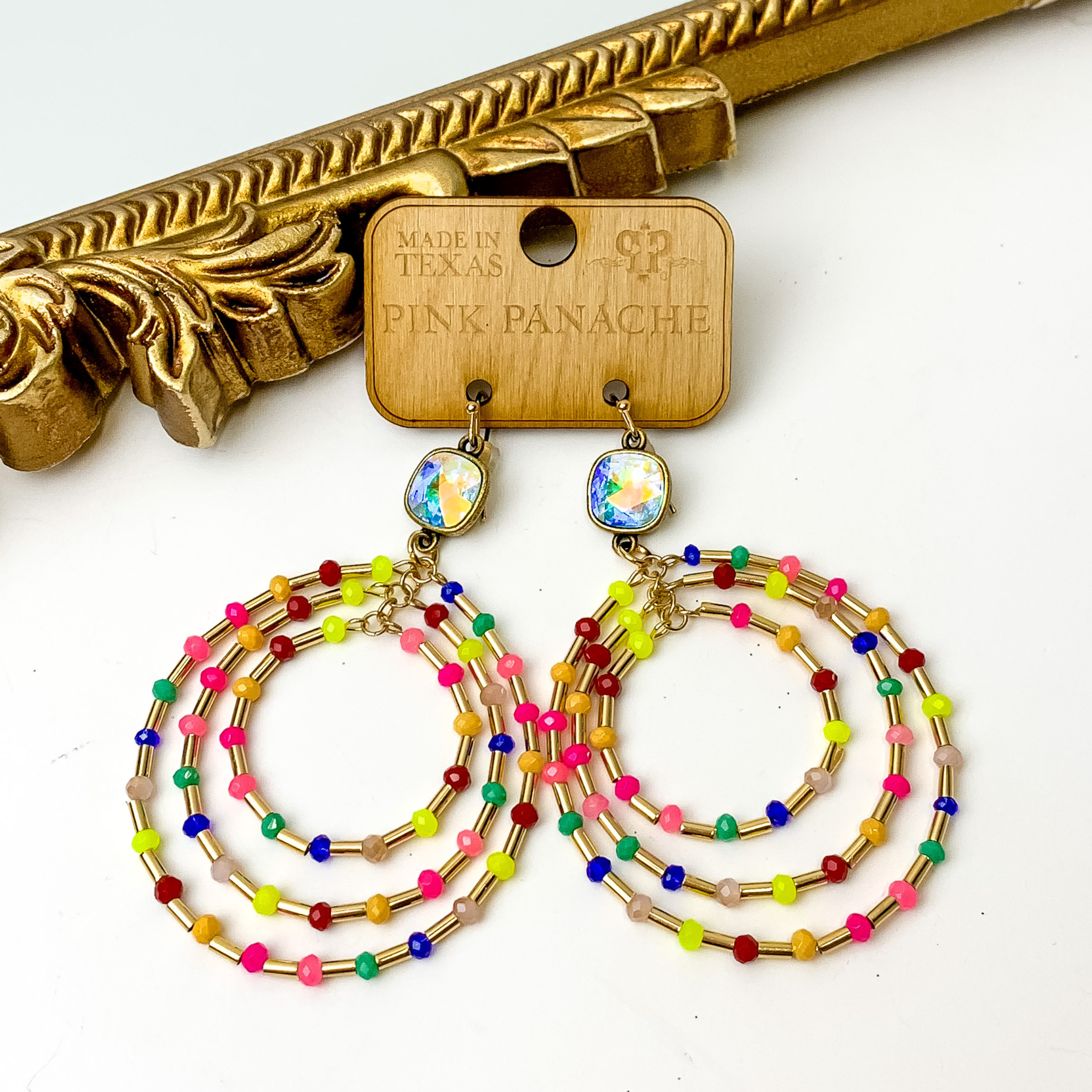 Ab cushion cut crystal with three beaded hoops. The hoop include gold and multicolor beads. These earrings are pictured on a white background with a gold mirror in the top left corner. 