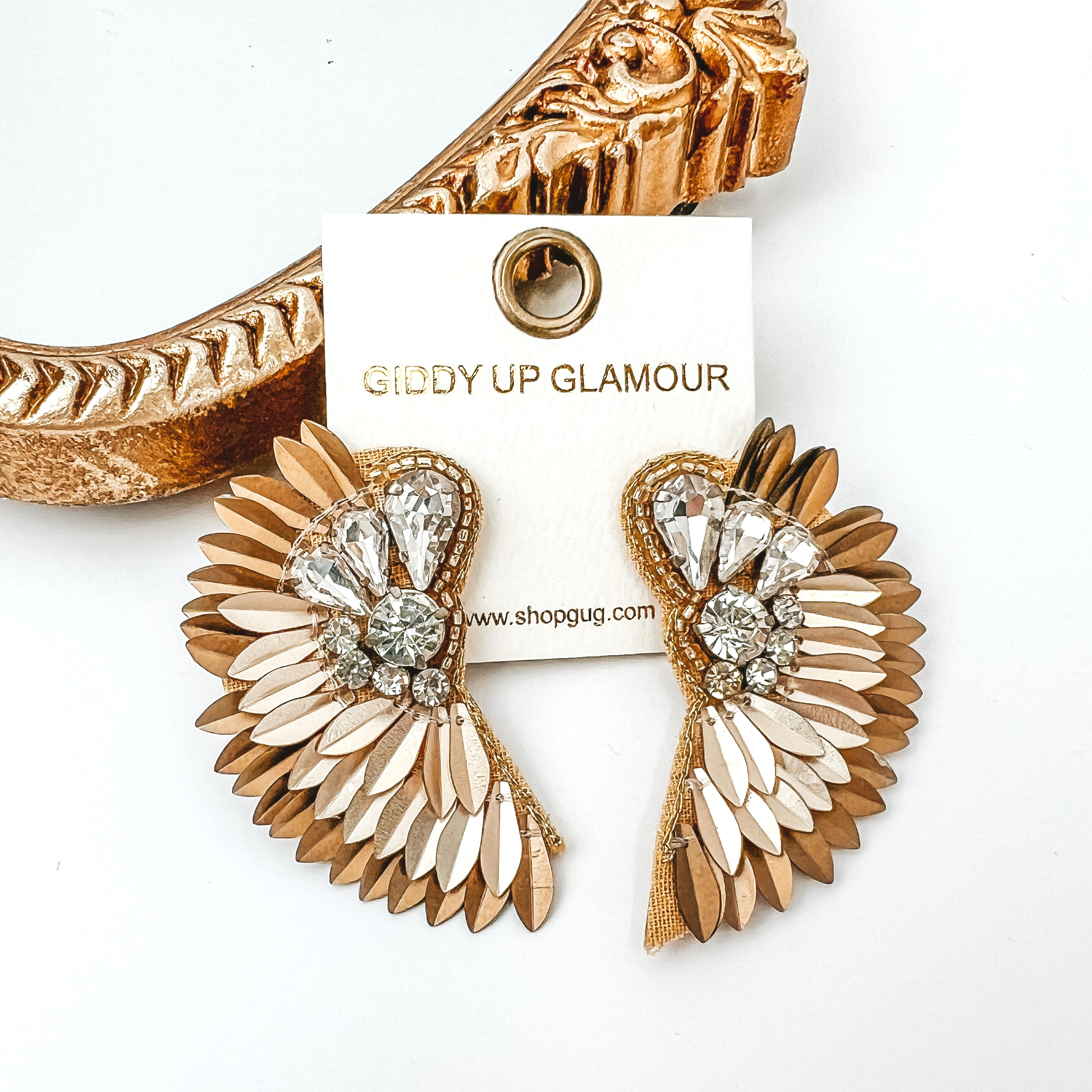 Gold and light gold, sequin feather earrings with a clear crystal design. These earrings are pictured on a white background with a gold mirror in the top left corner. 