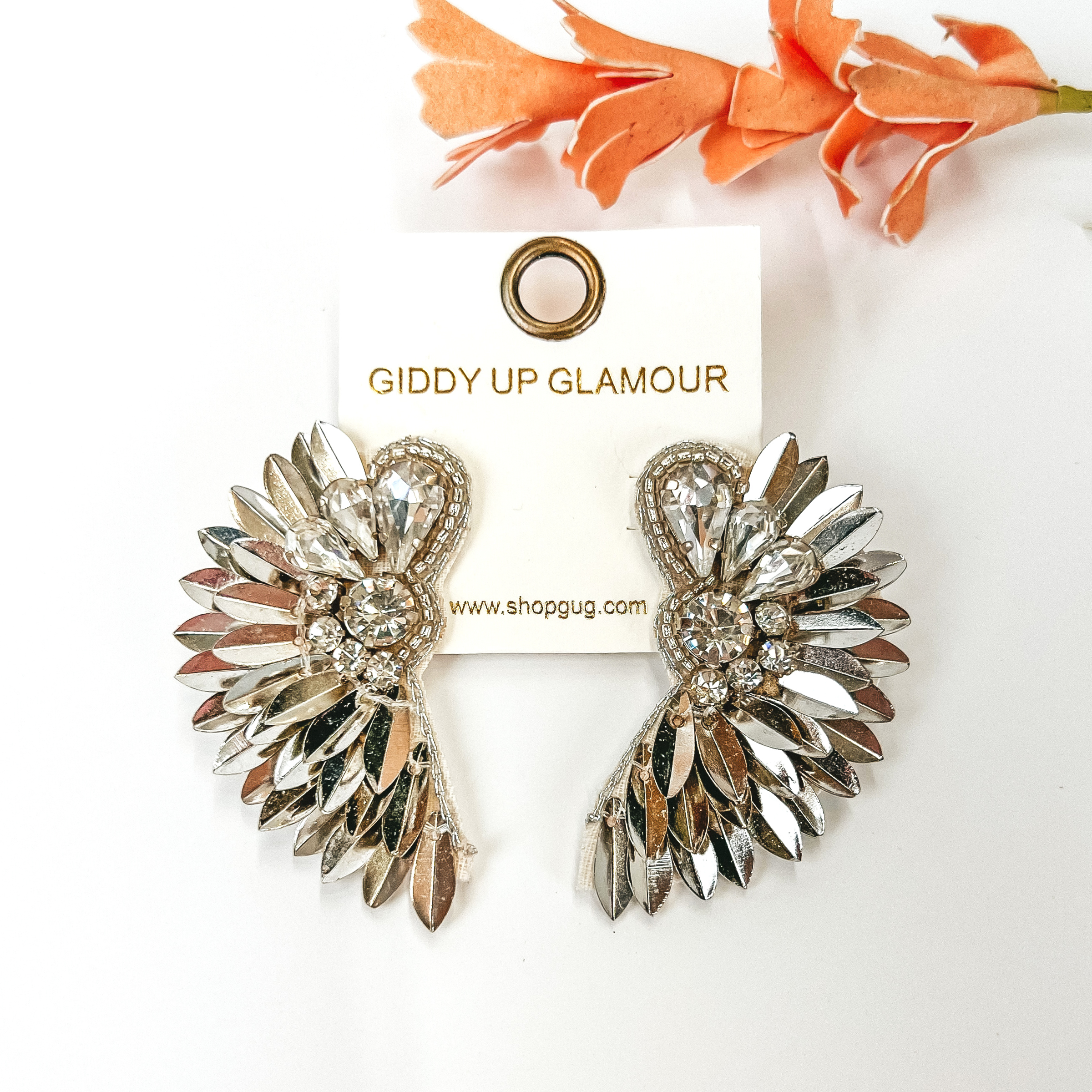 Silver, sequin feather earrings with a clear crystal design. These earrings are pictured on a white background with an orange flower above the earrings. 