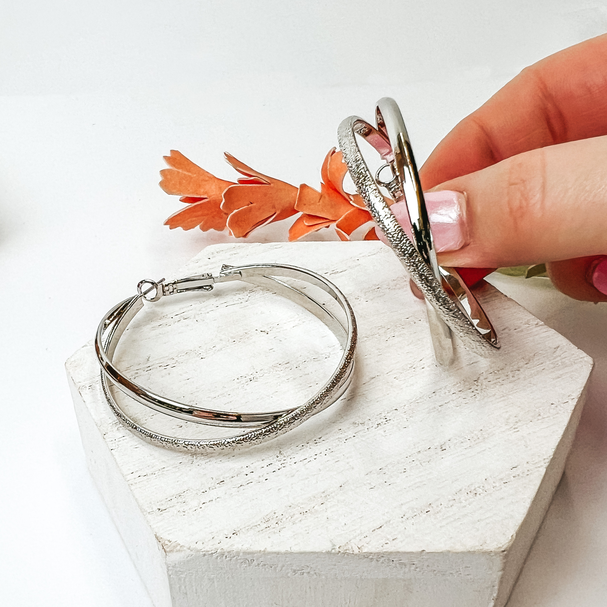 Double hoop earrings in silver. One hoop is silver and the other one is a texturized silver. One hoop is pictured on a white block while the other one is being held by a hand all on a white background. 