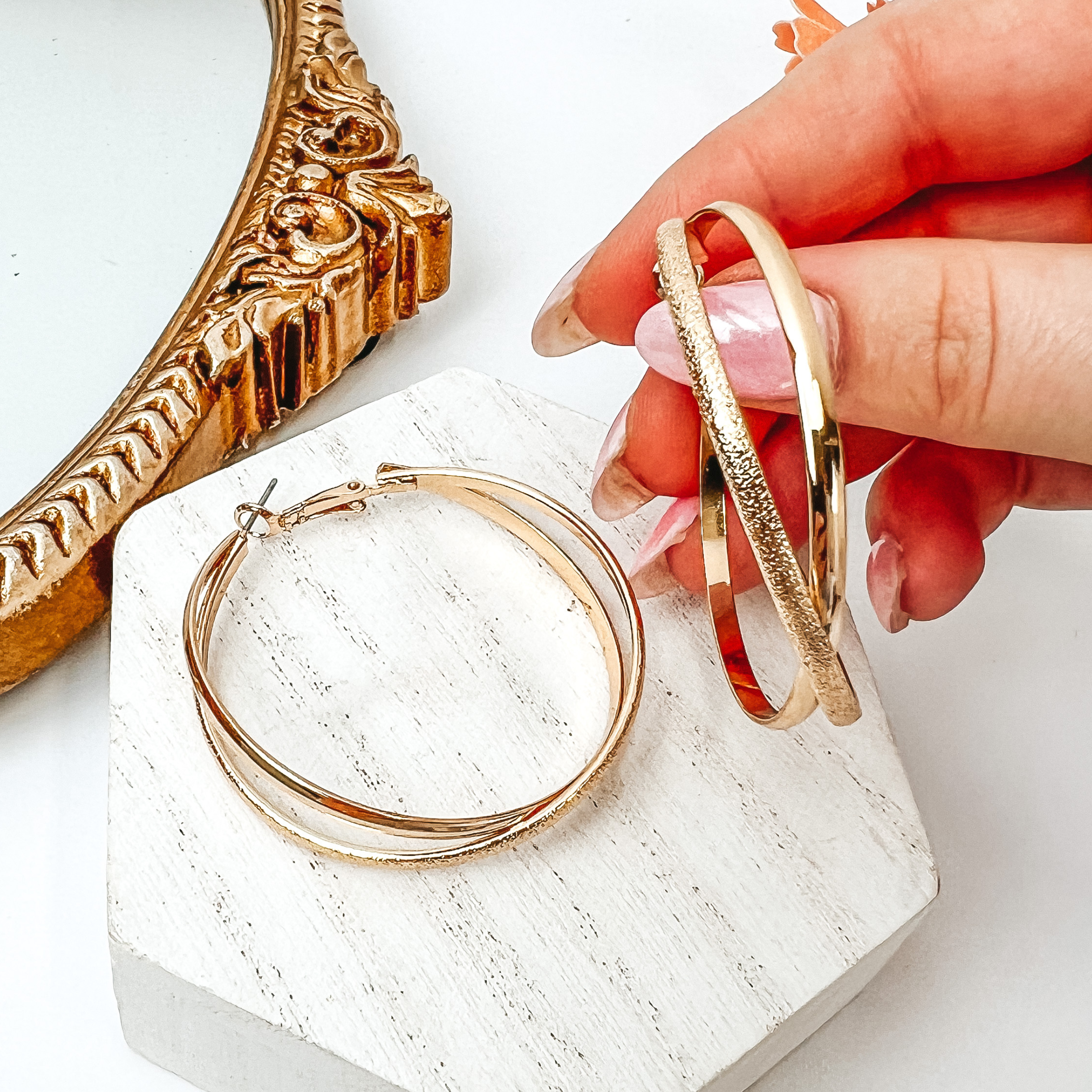 Double hoop earrings in gold. One hoop is gold and the other one is a texturized gold. One hoop is pictured on a white block while the other one is being held by a hand all on a white background. 