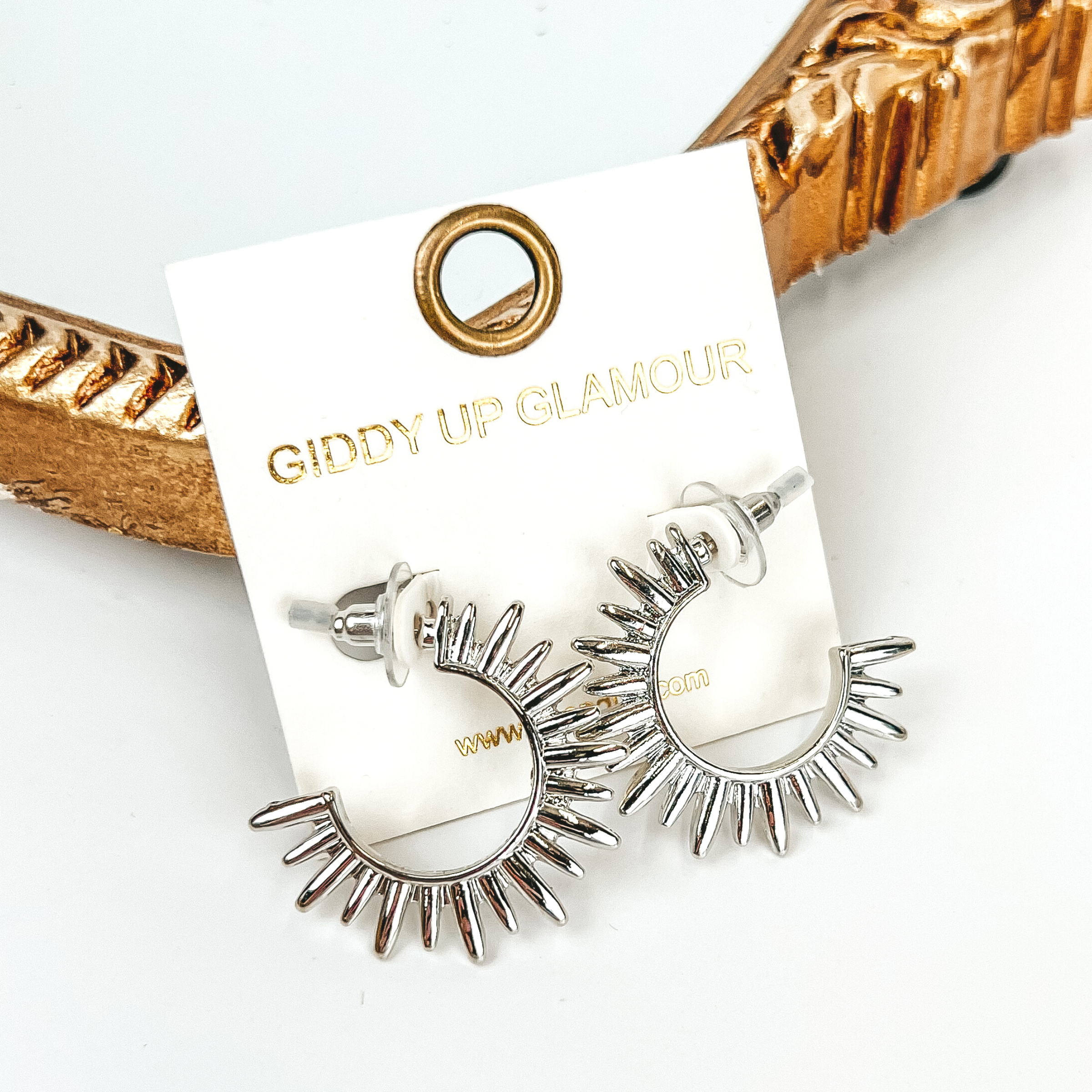 Silver sunburst hoop earrings. These earrings are pictured on a white background with a gold mirror behind the earrings. 