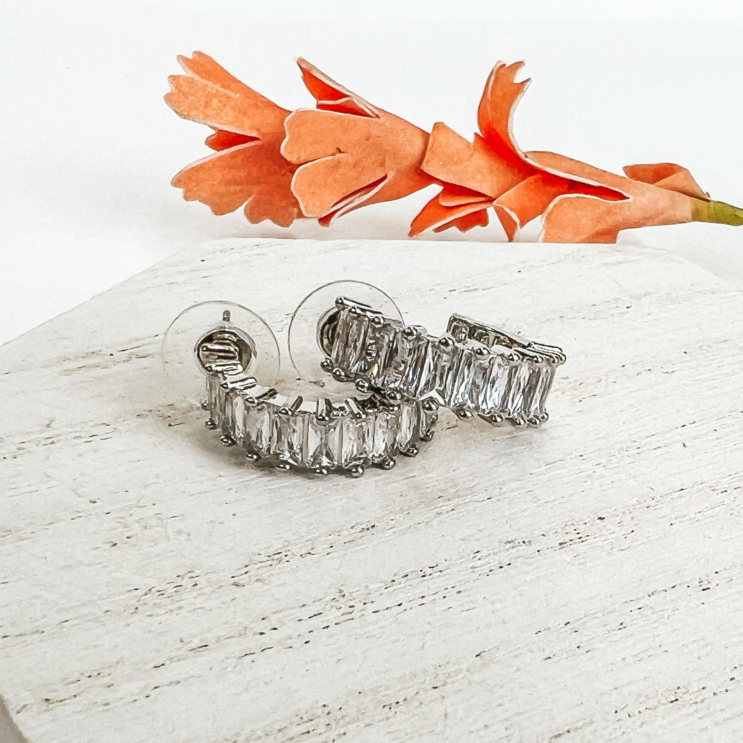 Silver hoop earrings with rectangle clear crystals. These earrings are pictured on a white background with an orange flower behind them. 