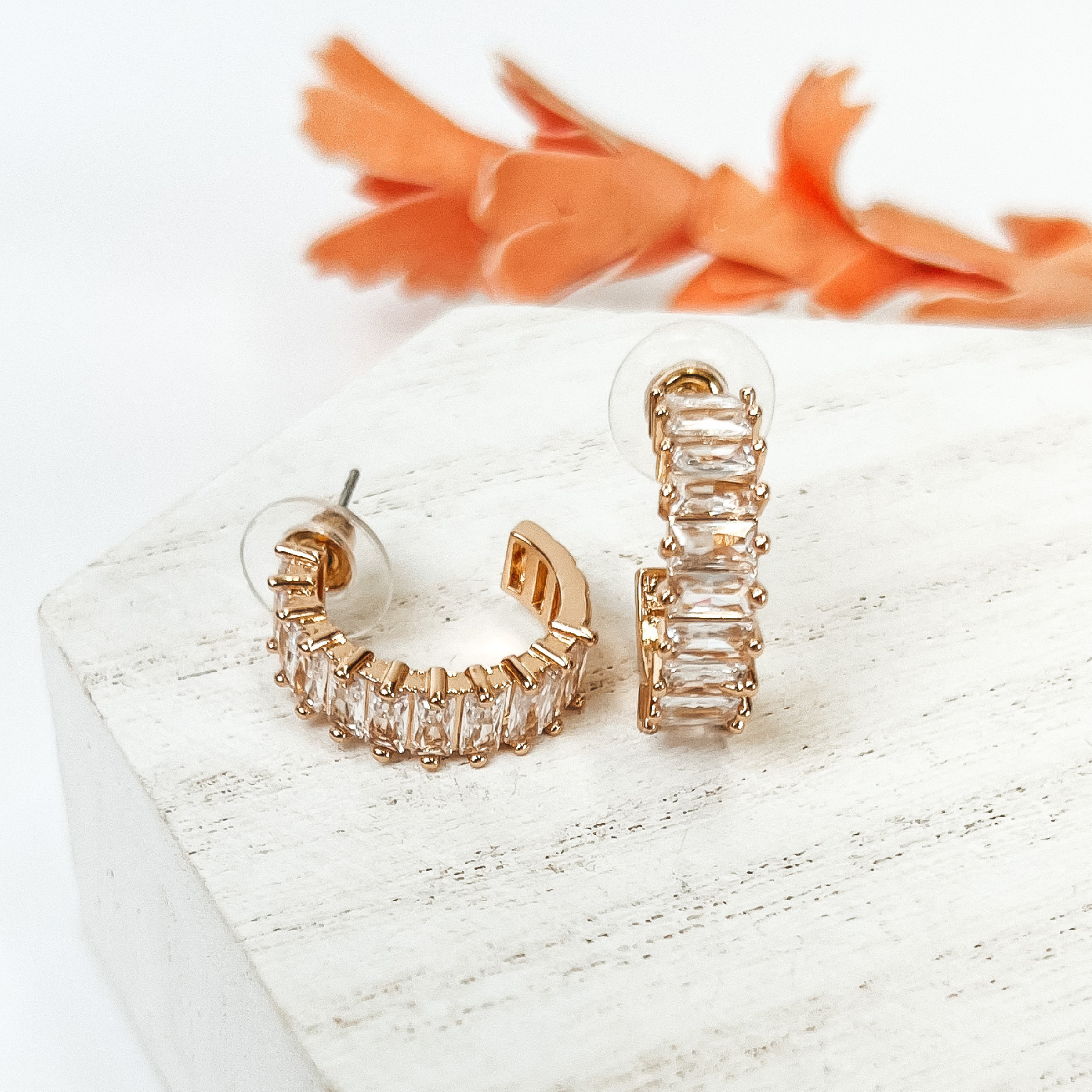 Gold hoop earrings with rectangle clear crystals. These earrings are pictured on a white background with an orange flower behind them. 