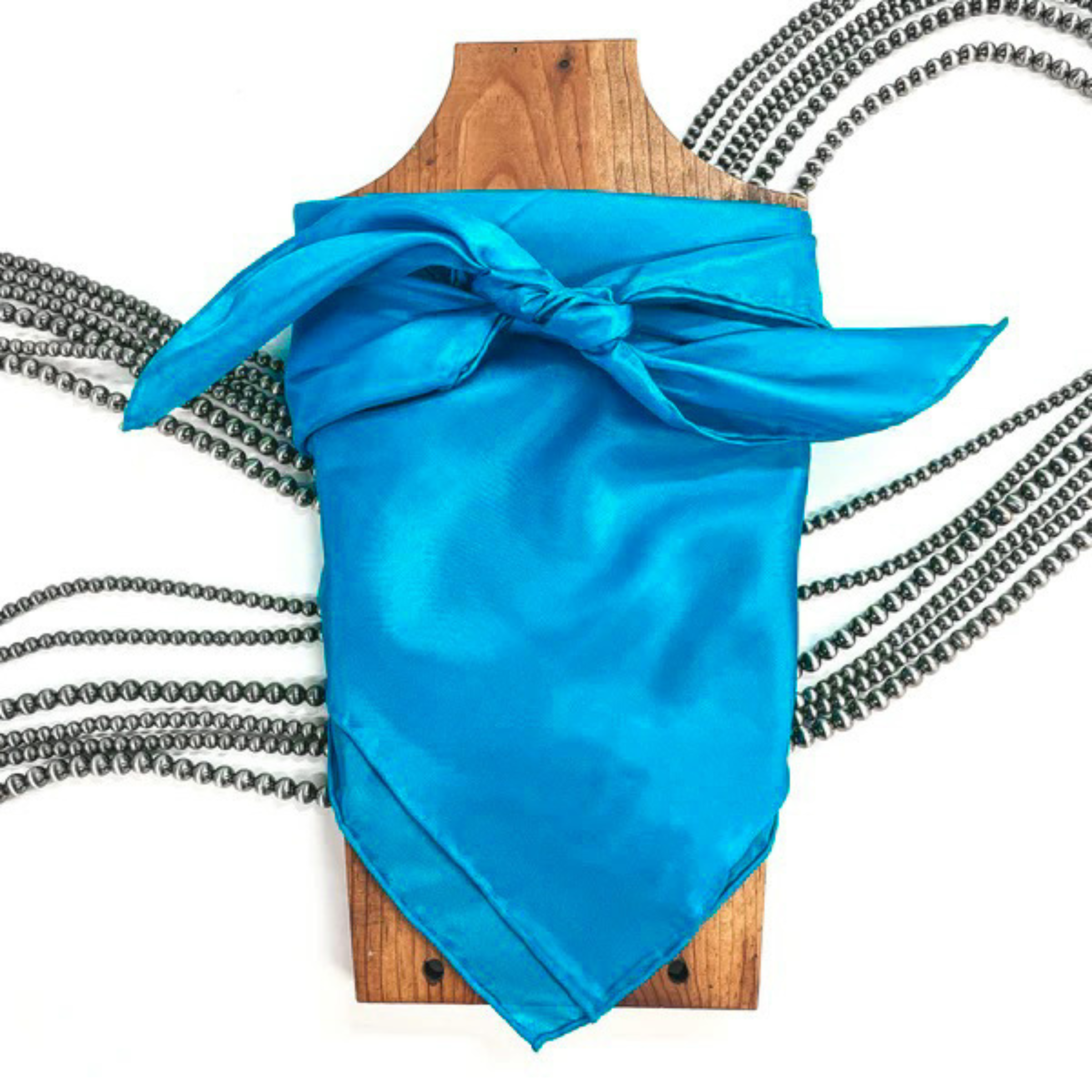 This is a turquoise silk wild rag, this wild rag is placed on a brown necklace board. This wild rag is taken on a white background and with silver Navajo pearls in the back as decor.