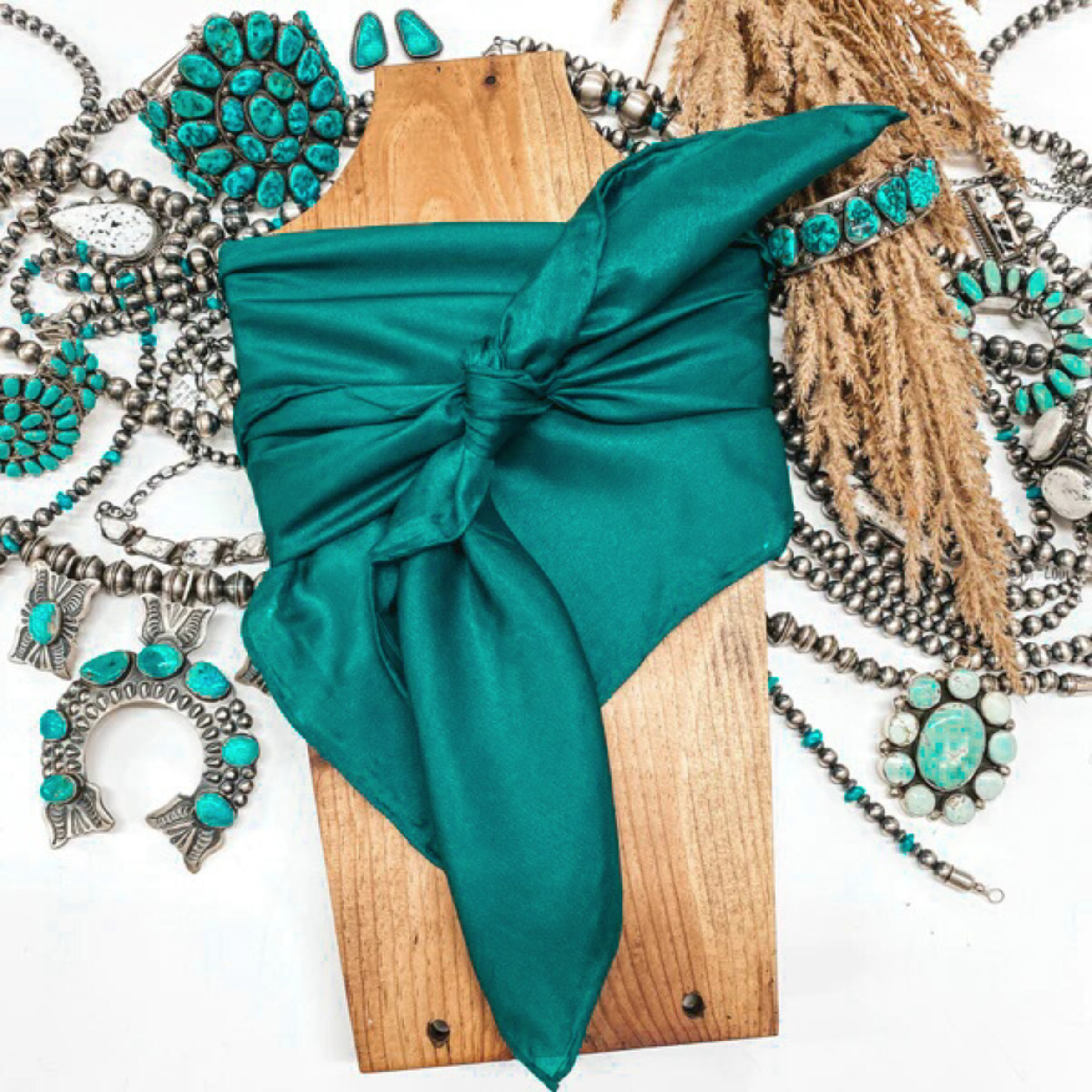 This is a teal silk wild rag, this wild rag is pictured wrapped around a brown necklace board. This wild rag is pictured on a white background and with silver and turquoise Navajo jewelry behind it as decoration.