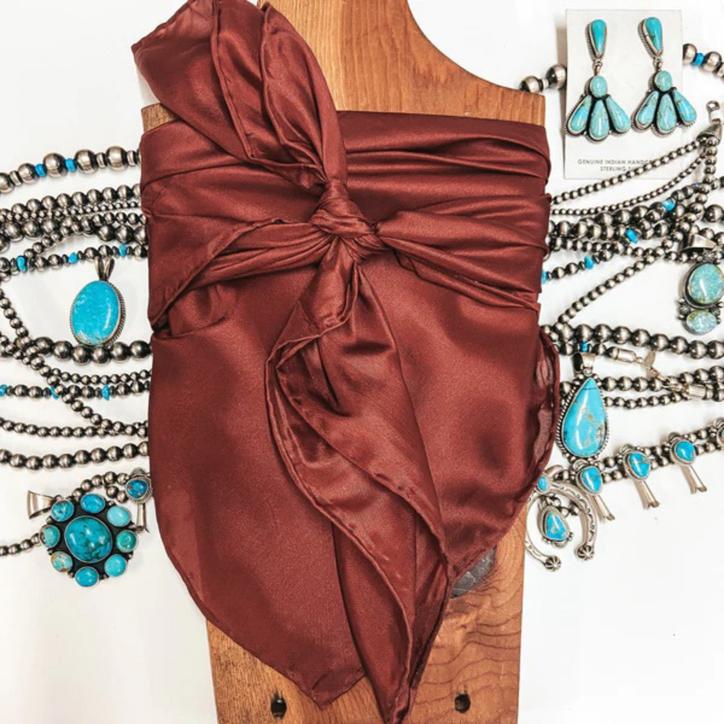 A solid color wild rag tied around a wooden display. Pictured on white background with turquoise jewelry.