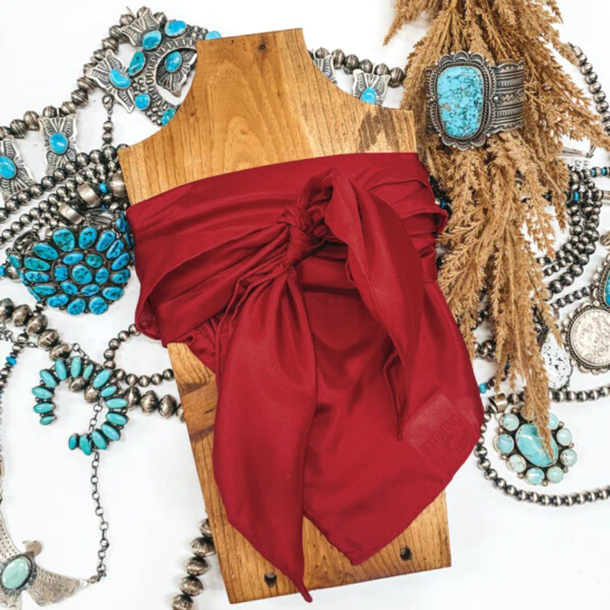 This is a deep red silk wild rag, this wild rag is pictured wrapped around a brown necklace board. This wild rag is pictured on a white background and with silver and turquoise Navajo jewelry behind it as decoration.
