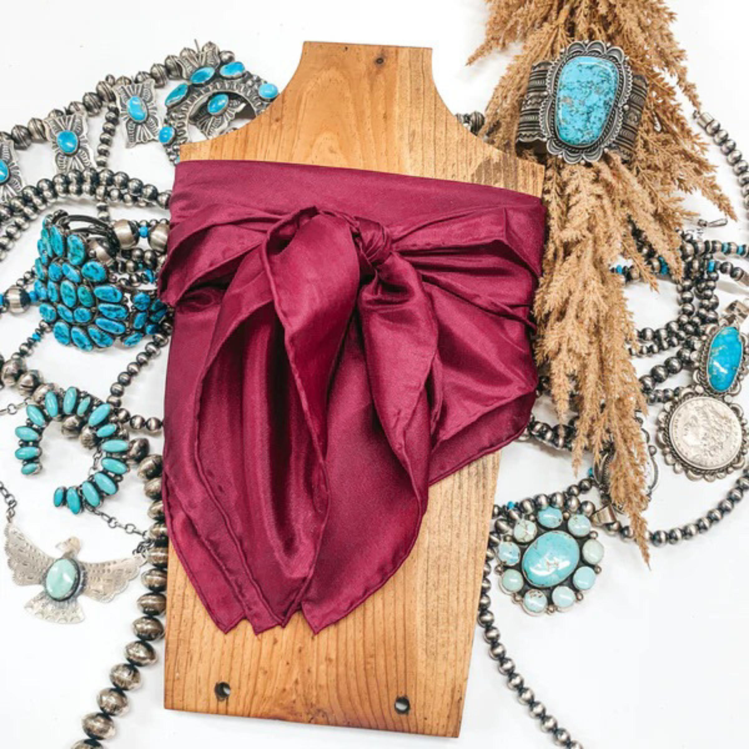 This is a maroon silk wild rag, this wild rag is pictured wrapped around a brown necklace board. This wild rag is pictured on a white background and with silver and turquoise Navajo jewelry behind it as decoration.