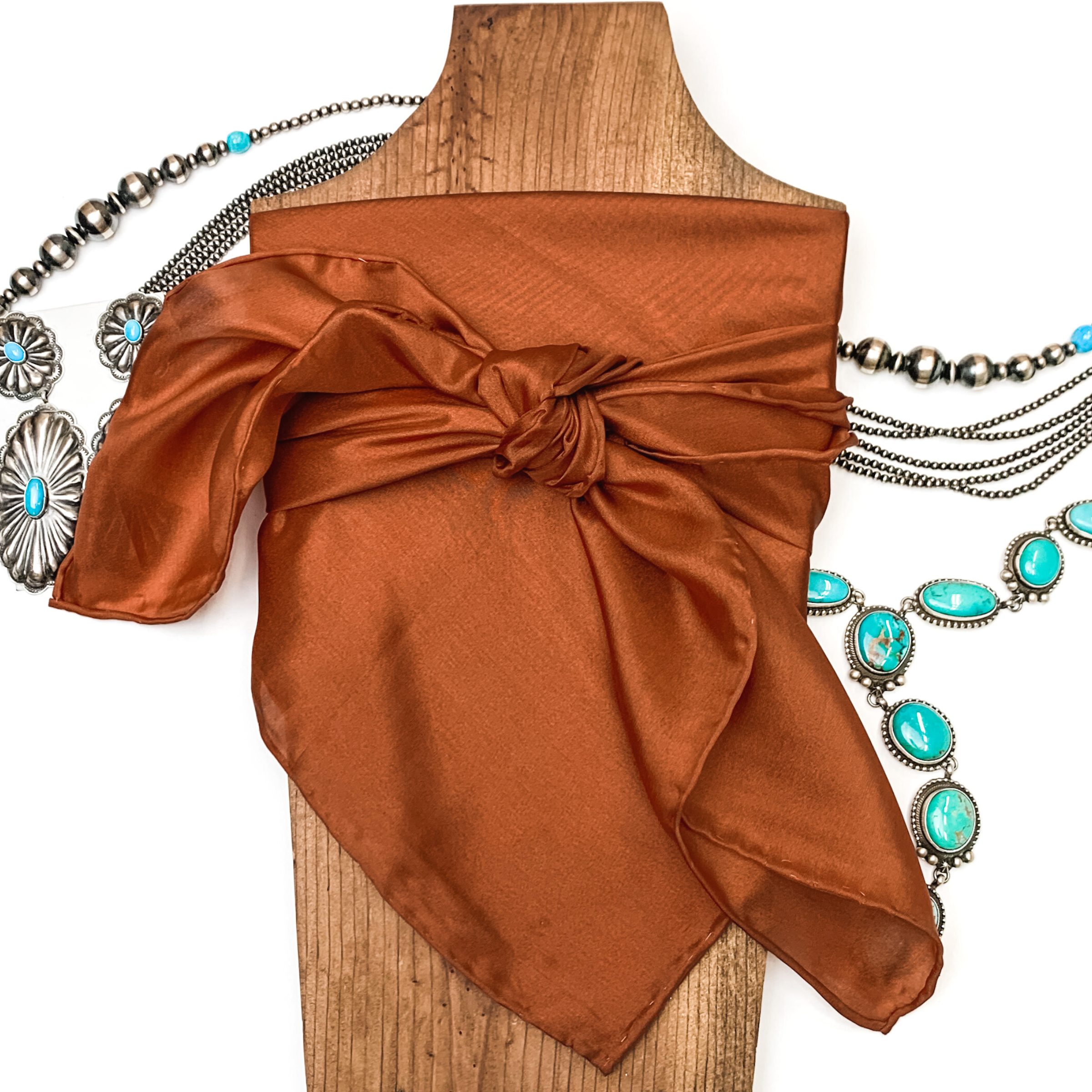 This is a copper silk wild rag, this wild rag is pictured wrapped around a brown necklace board. This wild rag is pictured on a white background and with silver and turquoise Navajo jewelry behind it as decoration.