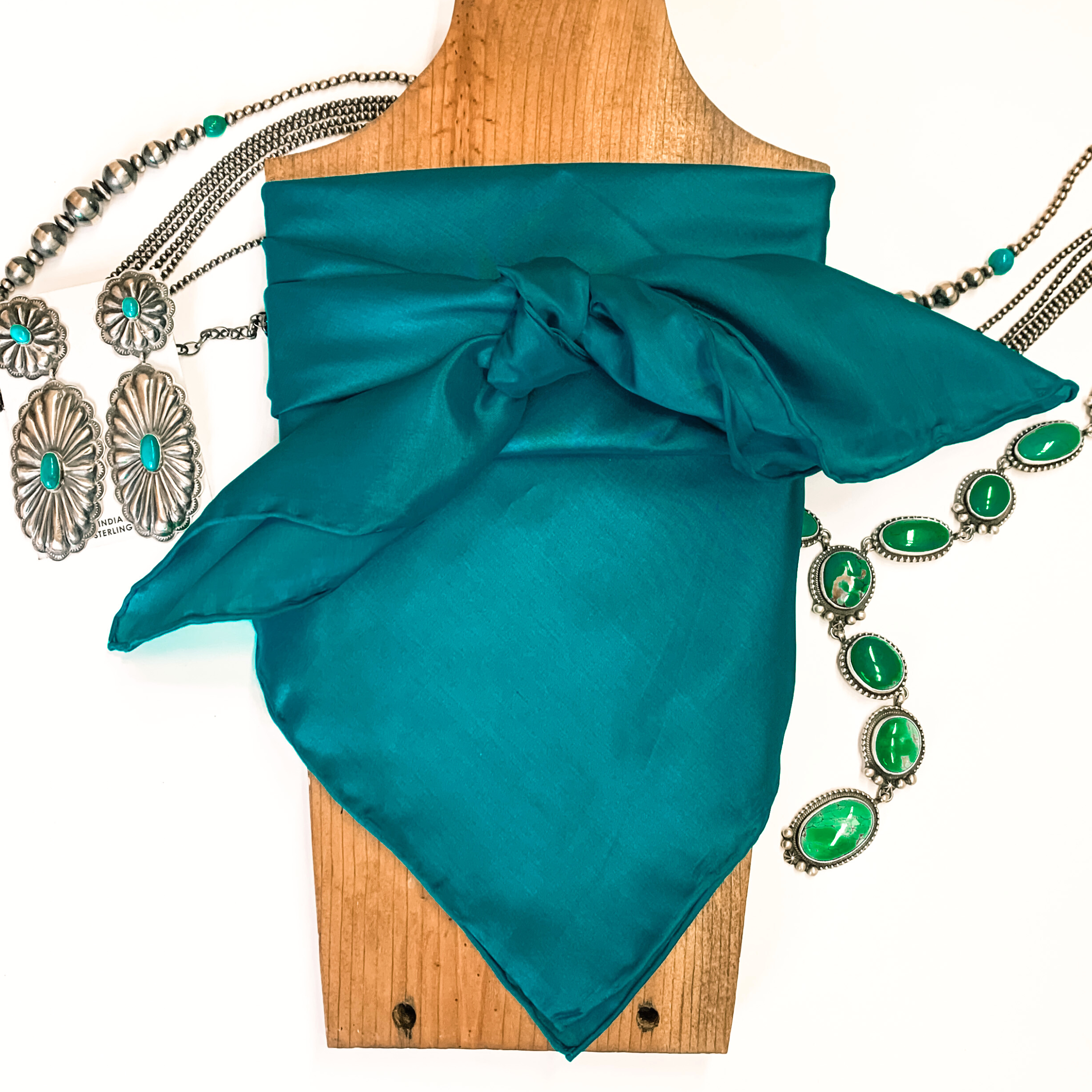 This is a aqua silk wild rag, this wild rag is pictured wrapped around a brown necklace board. This wild rag is pictured on a white background and with silver and turquoise Navajo jewelry behind it as decoration.