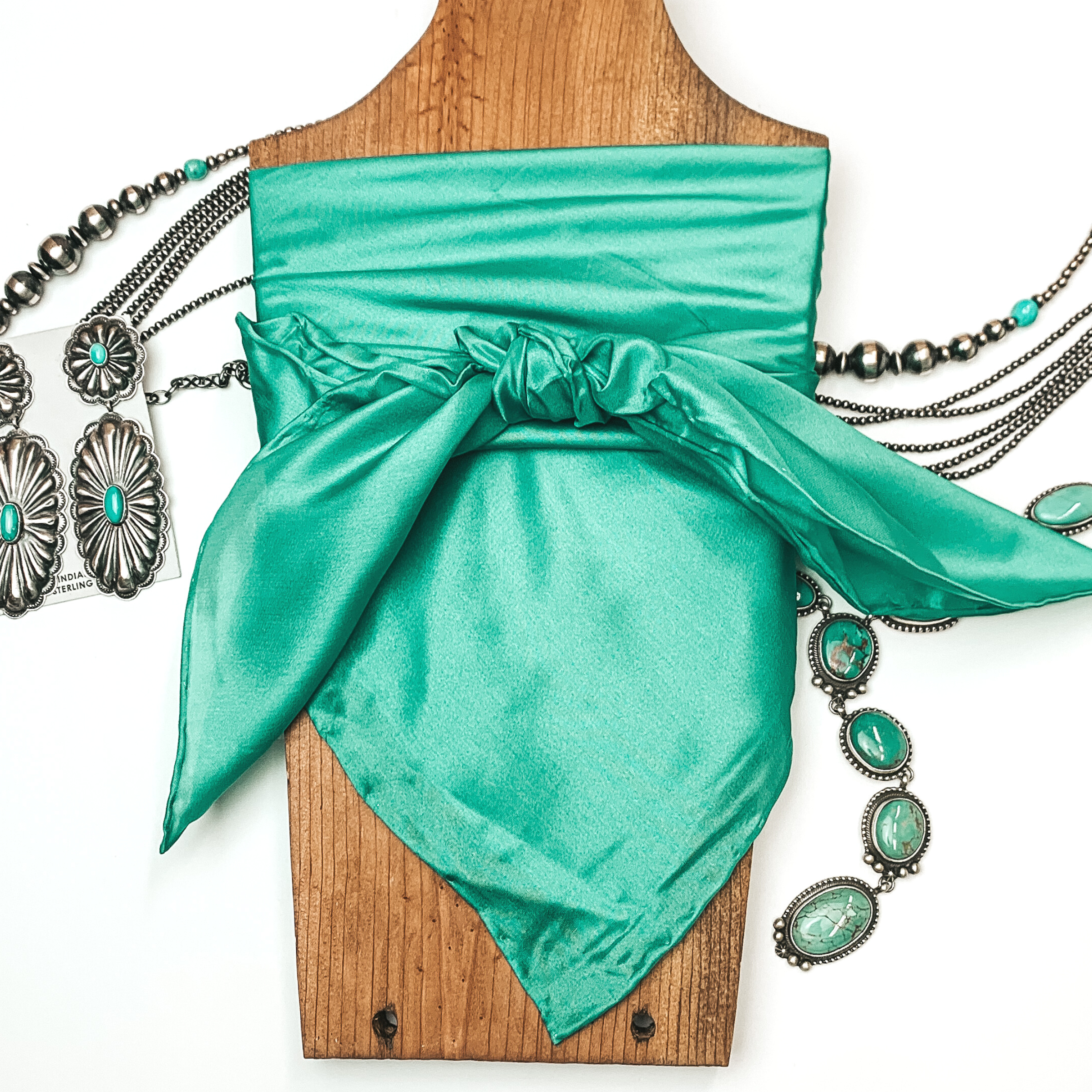 This is a emerald green silk wild rag, this wild rag is pictured wrapped around a brown necklace board. This wild rag is pictured on a white background and with silver and turquoise Navajo jewelry behind it as decoration.