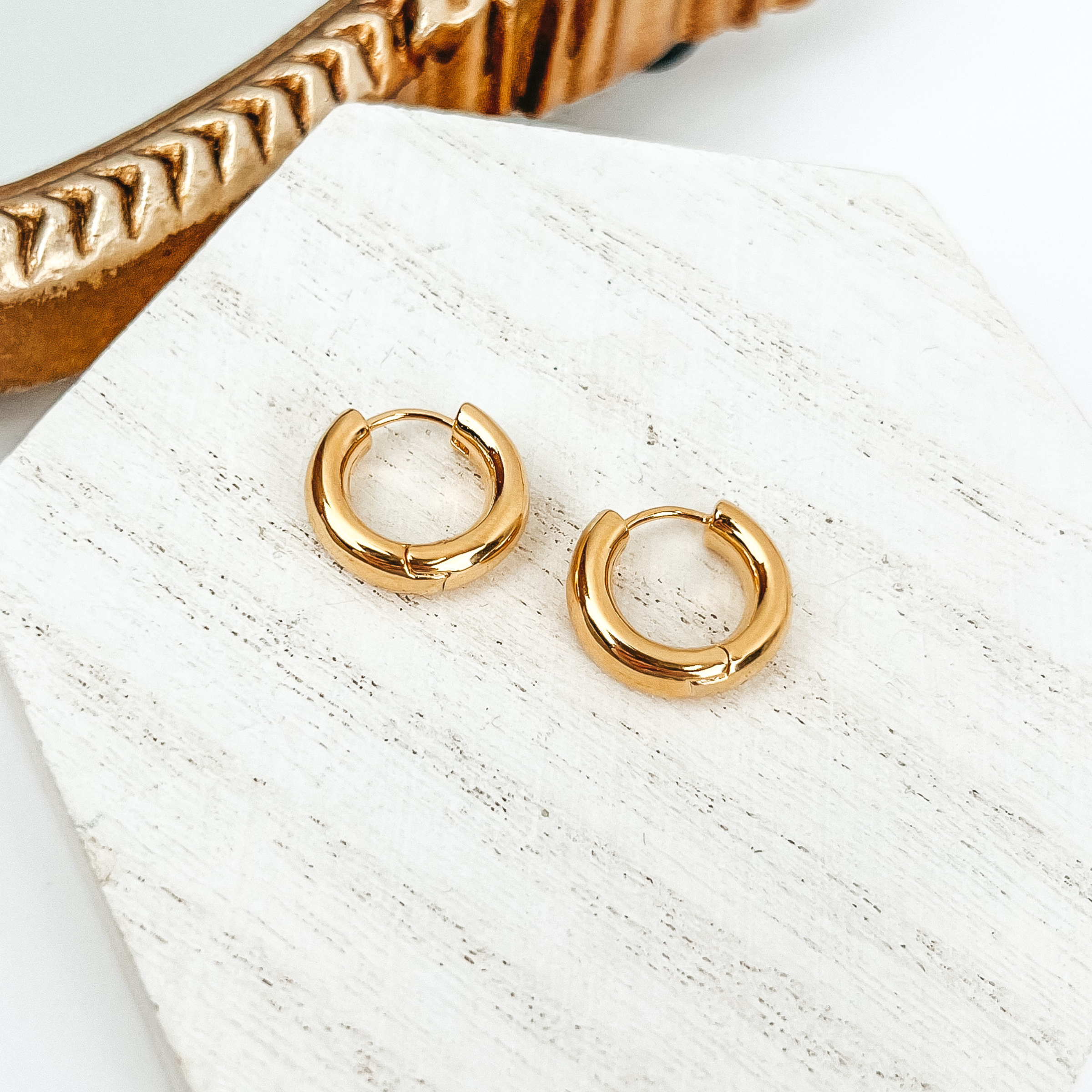 Gold hoop earrings pictured on a white block on a white background. There is also a gold mirror in the top left corner.