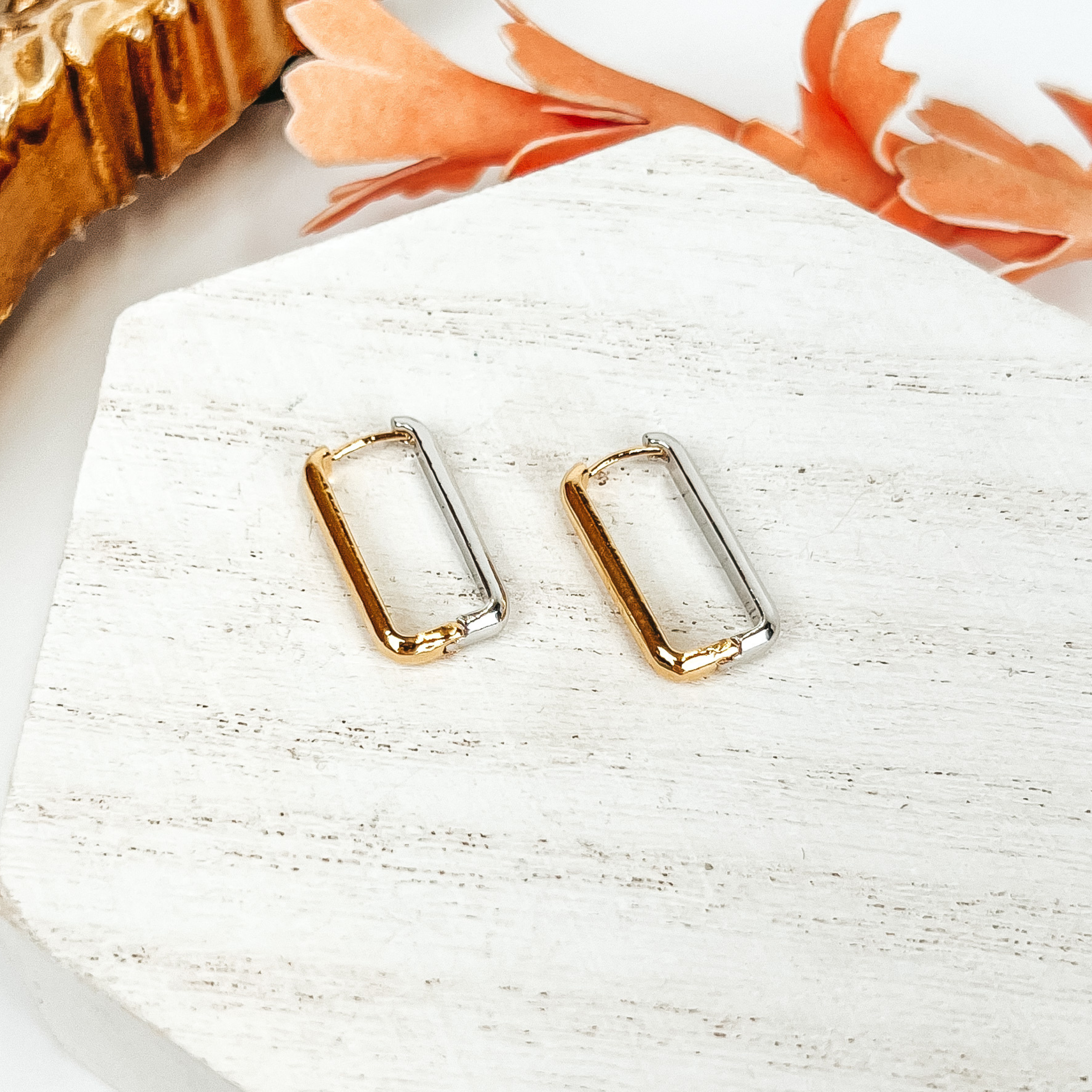 Half gold and half silver rectangle hoop earrings pictured on a white block on a white background. There is also a gold mirror in the top left corner.