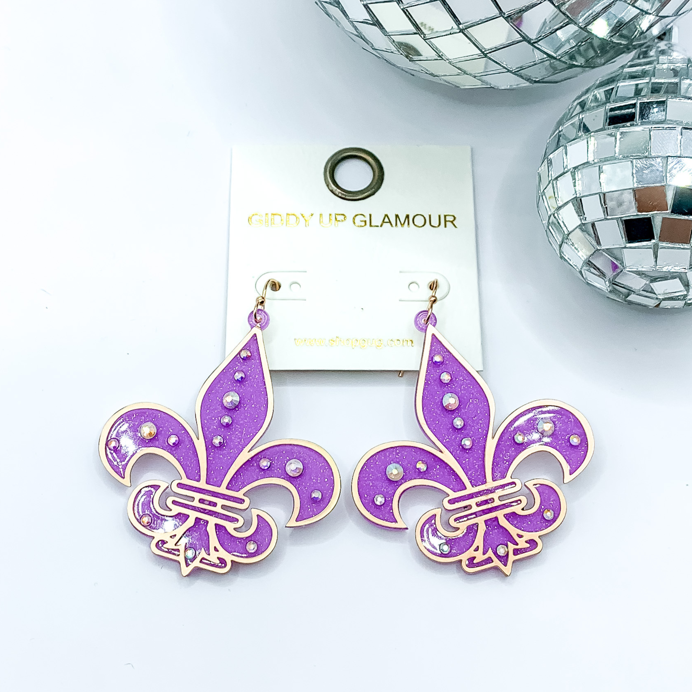 Purple glitter fleur de lis dangle earrings. These earrings include a gold outline and ab crystals. These earrings are pictured on a white background with disco balls in the top right corner.