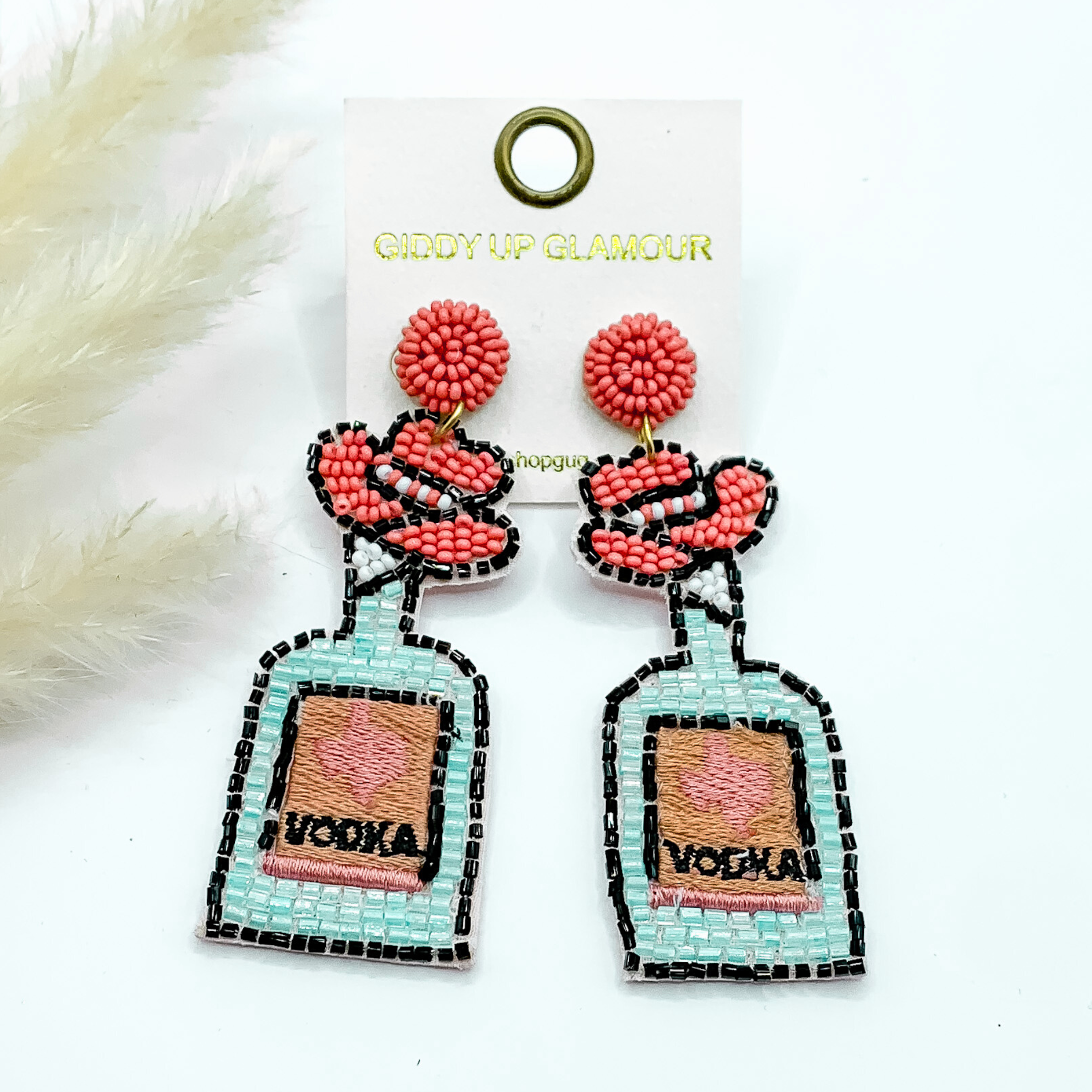 Pink beaded circle post earrings with beaded a pink cowboy hat and bottle pendant. the bottle has a black beaded outline and the word "VODKA" and a pink texas stitched in the center. These earrings are pictured on a white background. 