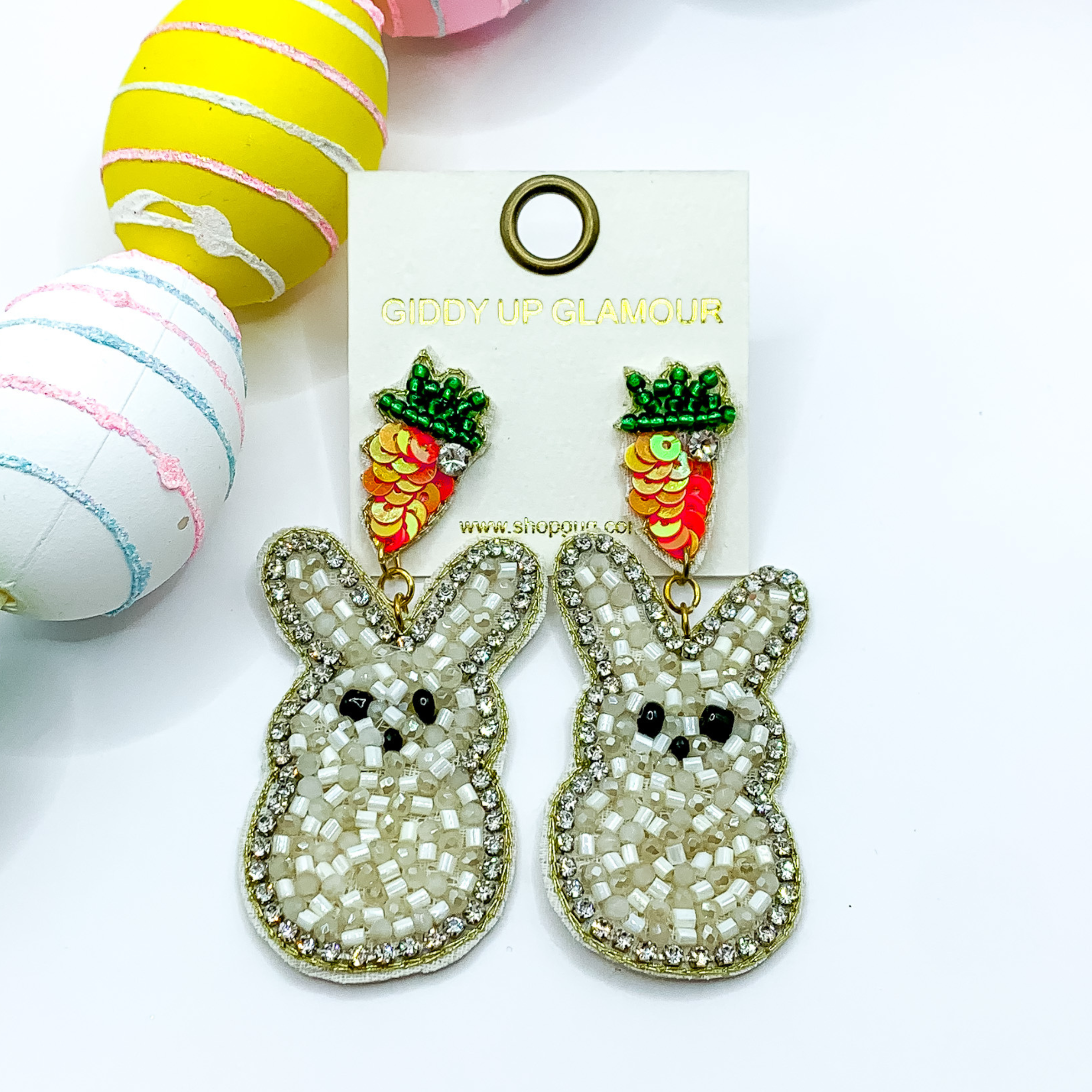Beaded, ivory bunny rabbit earrings on a carrot post with a clear crystal outline. Pictured on white background with colorful Easter eggs.