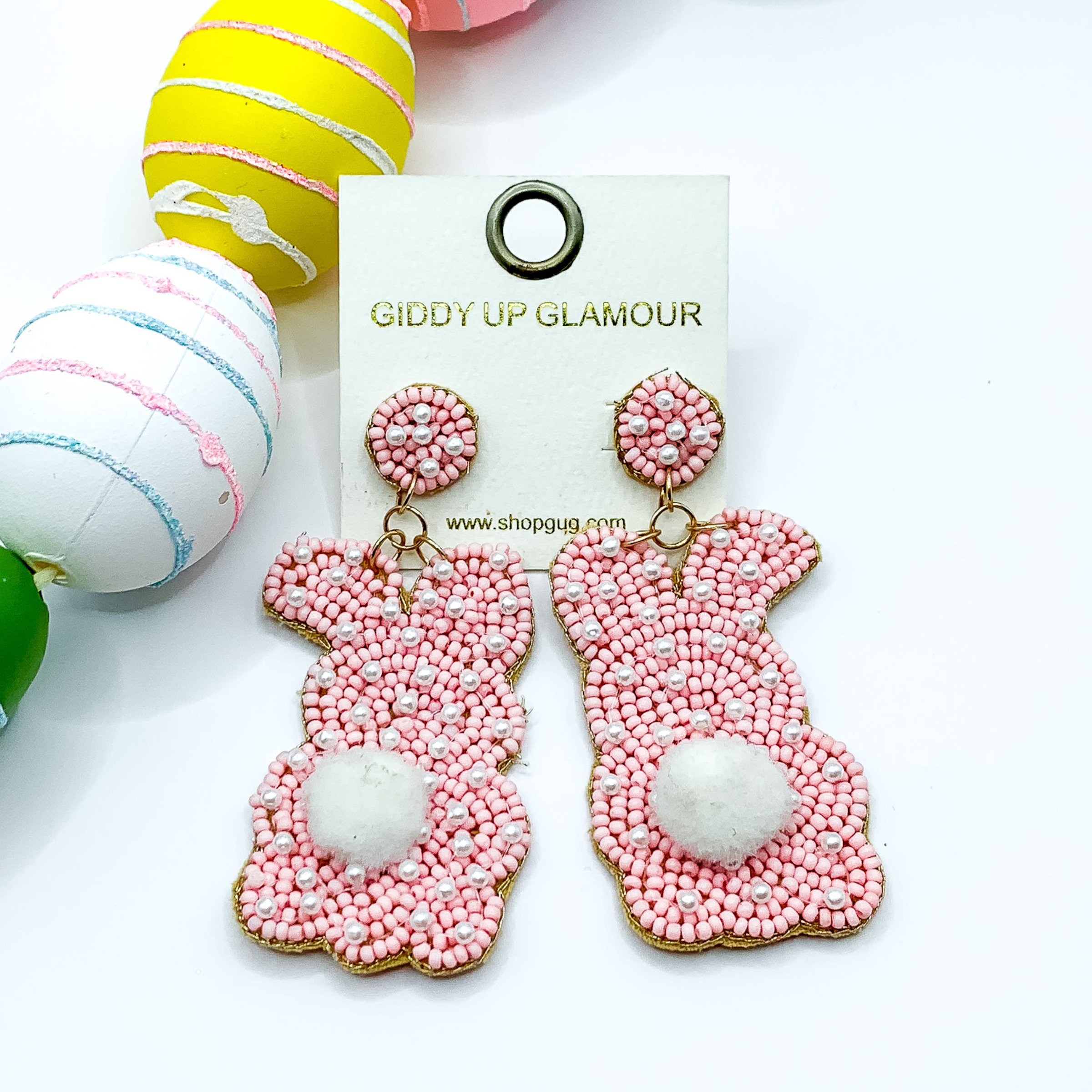 Beaded, baby pink bunny rabbit earrings with a white puff ball for a tail and a white beaded print. Pictured on white background with colorful Easter eggs.