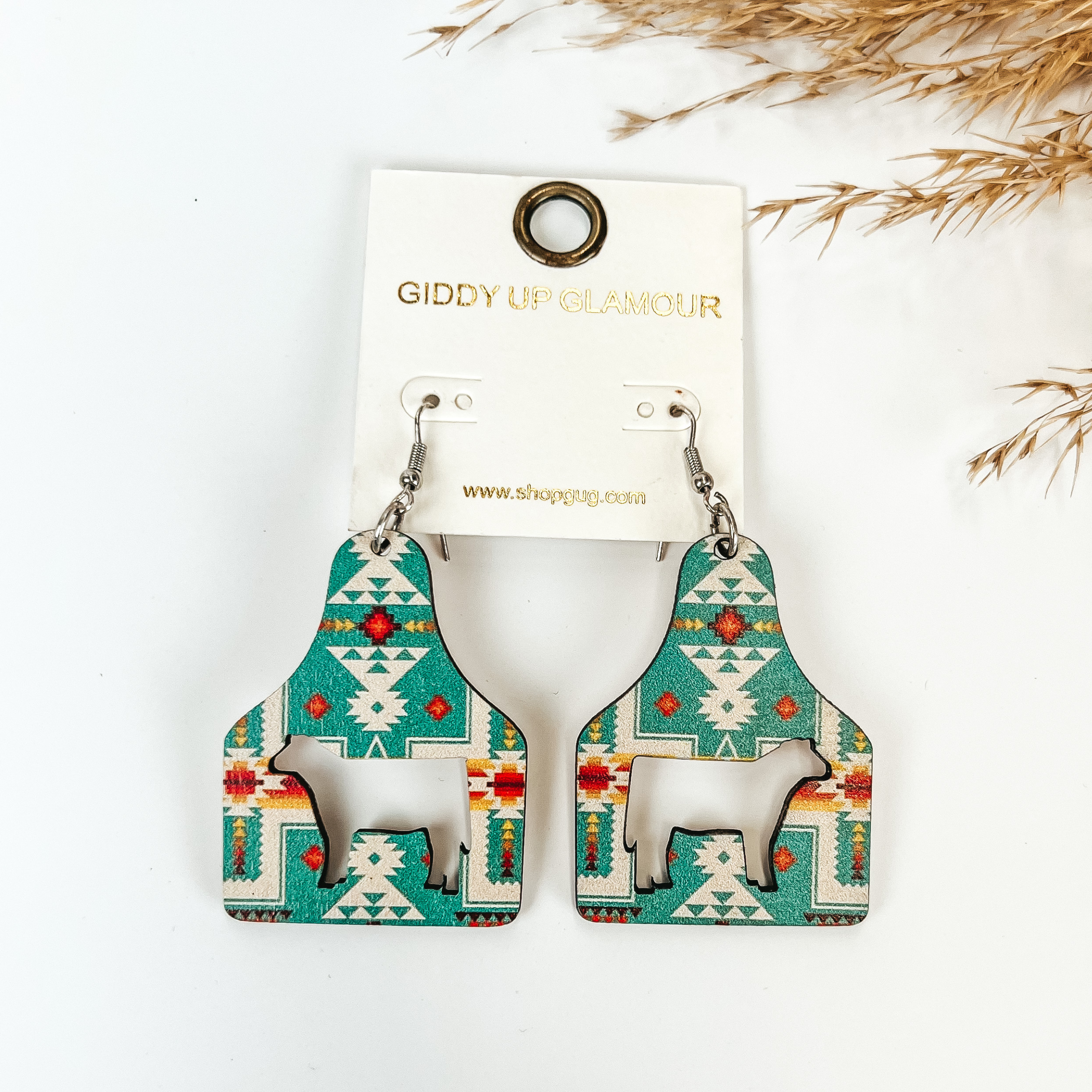 Cattle tag drop earrings with a turquoise aztec print. In the center of the earrings is a cutout of a cow. These earrings are pictured on a white background with tan pompous grass in the top left corner. 