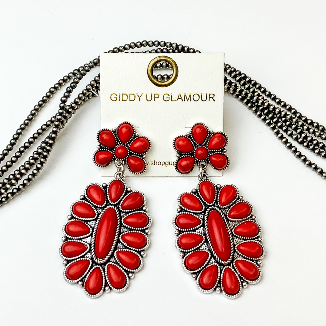 Silver, oval drop earrings with red stones. These earrings are pictured on a white background with silver beads behind the earrings. 