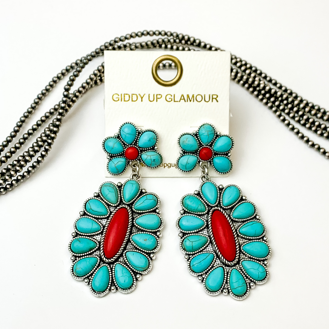 Silver, oval drop earrings with turquoise stones and red center stones. These earrings are pictured on a white background with silver beads behind the earrings. 