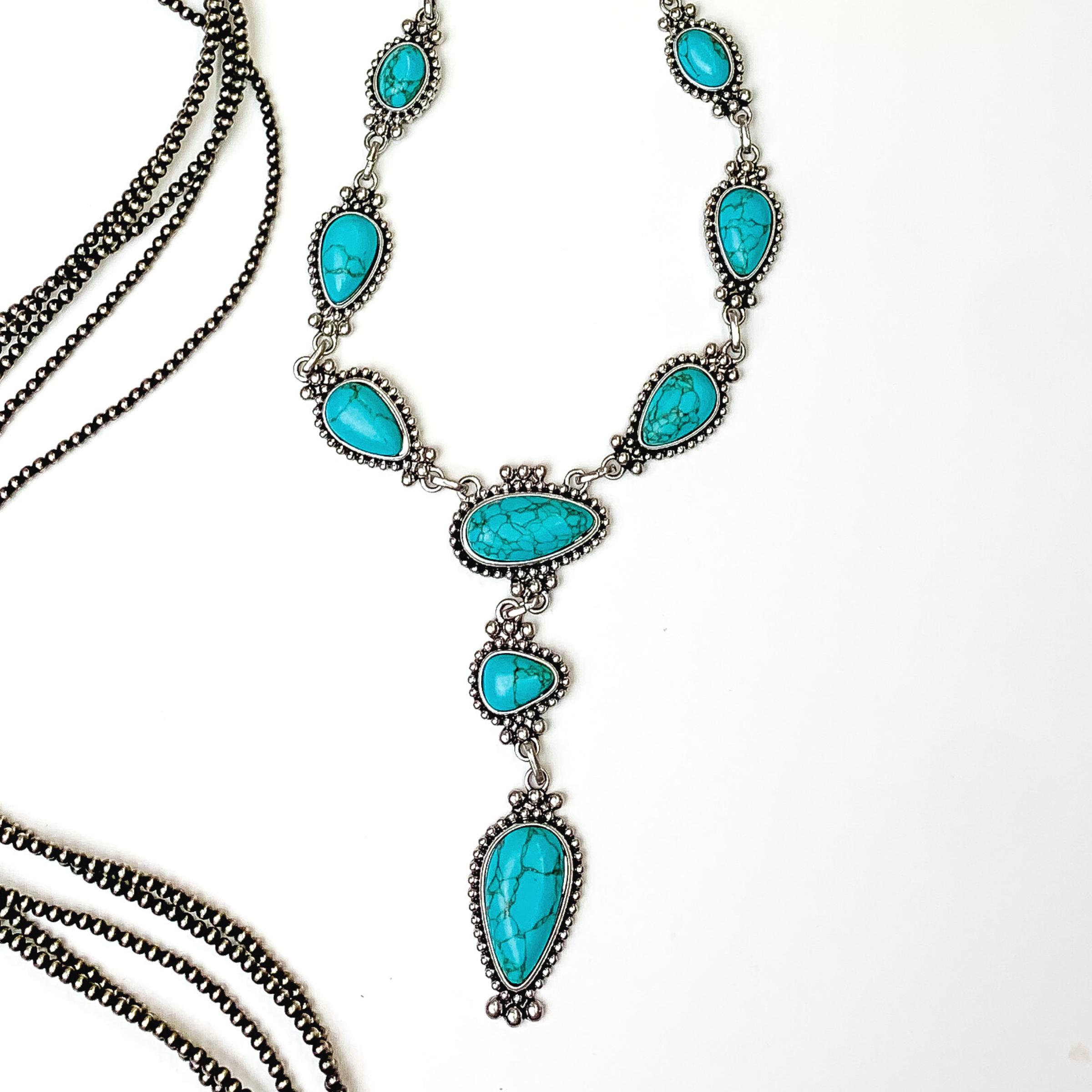 Silver lariat necklace with turquoise stones. Each stone has a silver beaded outline. This necklace is pictured on a white background with silver beads on the left side of the picture.