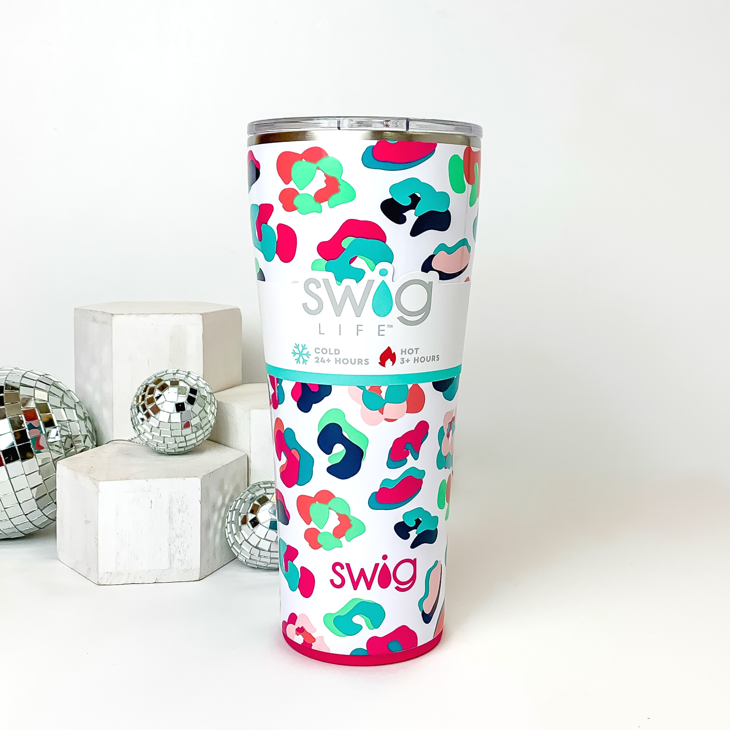White tumbler with a clear lid. This tumbler has a neon multicolored leopard print. This tumbler is pictured on a white background with white blocks and disco balls on the left side of the picture. 
