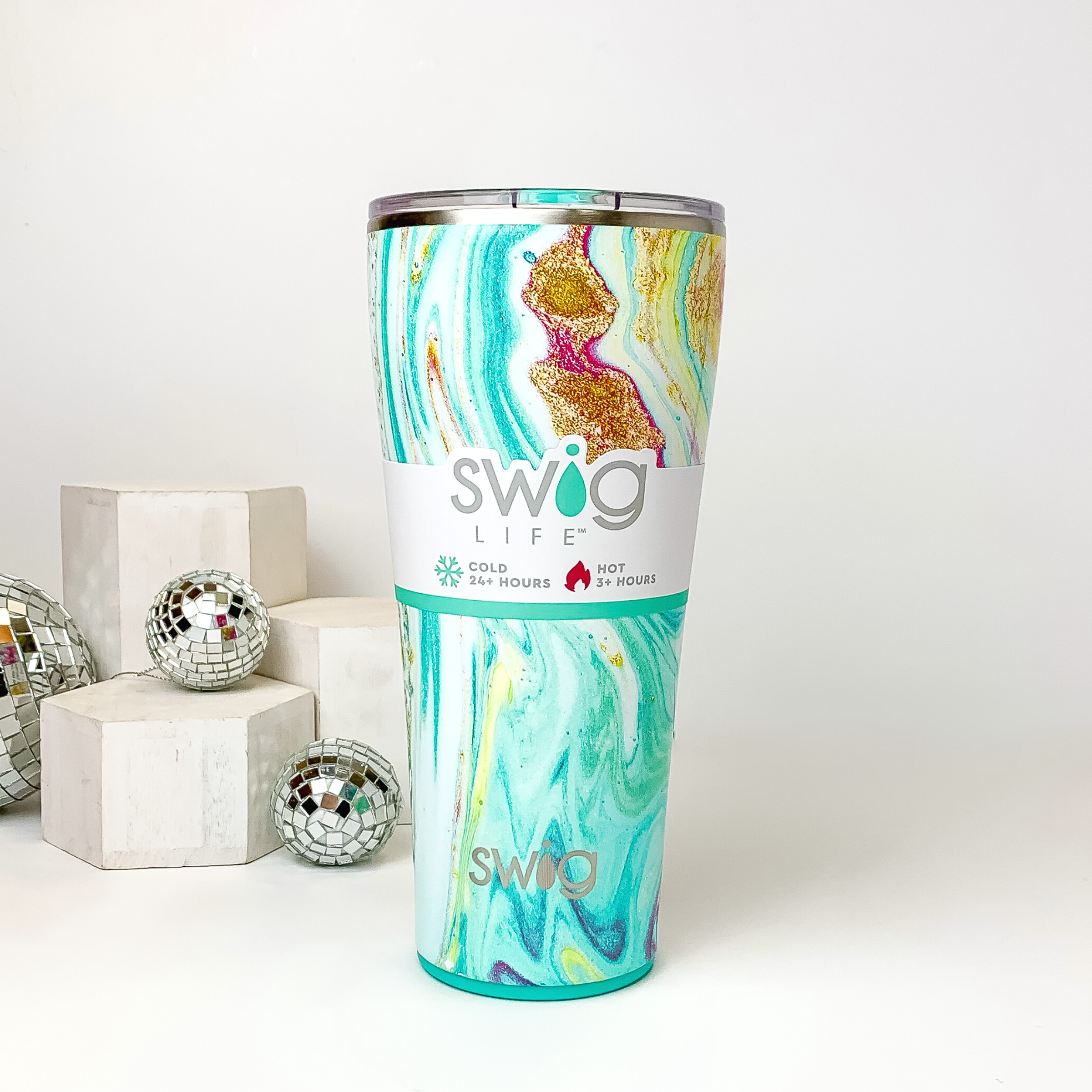 Aqua marble tumbler with a clear lid. This marble print includes shades of blue, pink, and gold glitter. This tumbler is pictured on a white background with white blocks and disco balls on the left side of the picture. 
