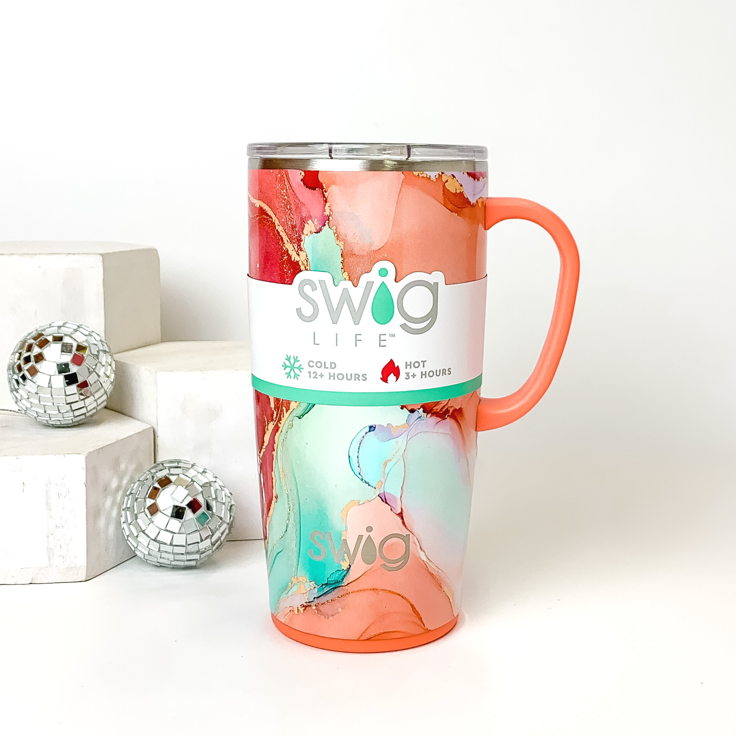 Pink and orange marble mug with a clear lid and orange handle. This marble print includes shades of orange, pink, aqua, and gold glitter. This mug is pictured on a white background with white blocks and disco balls on the left side of the picture. 