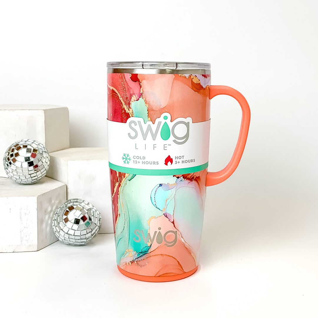 Pink and orange marble mug with a clear lid and orange handle. This marble print includes shades of orange, pink, aqua, and gold glitter. This mug is pictured on a white background with white blocks and disco balls on the left side of the picture. 