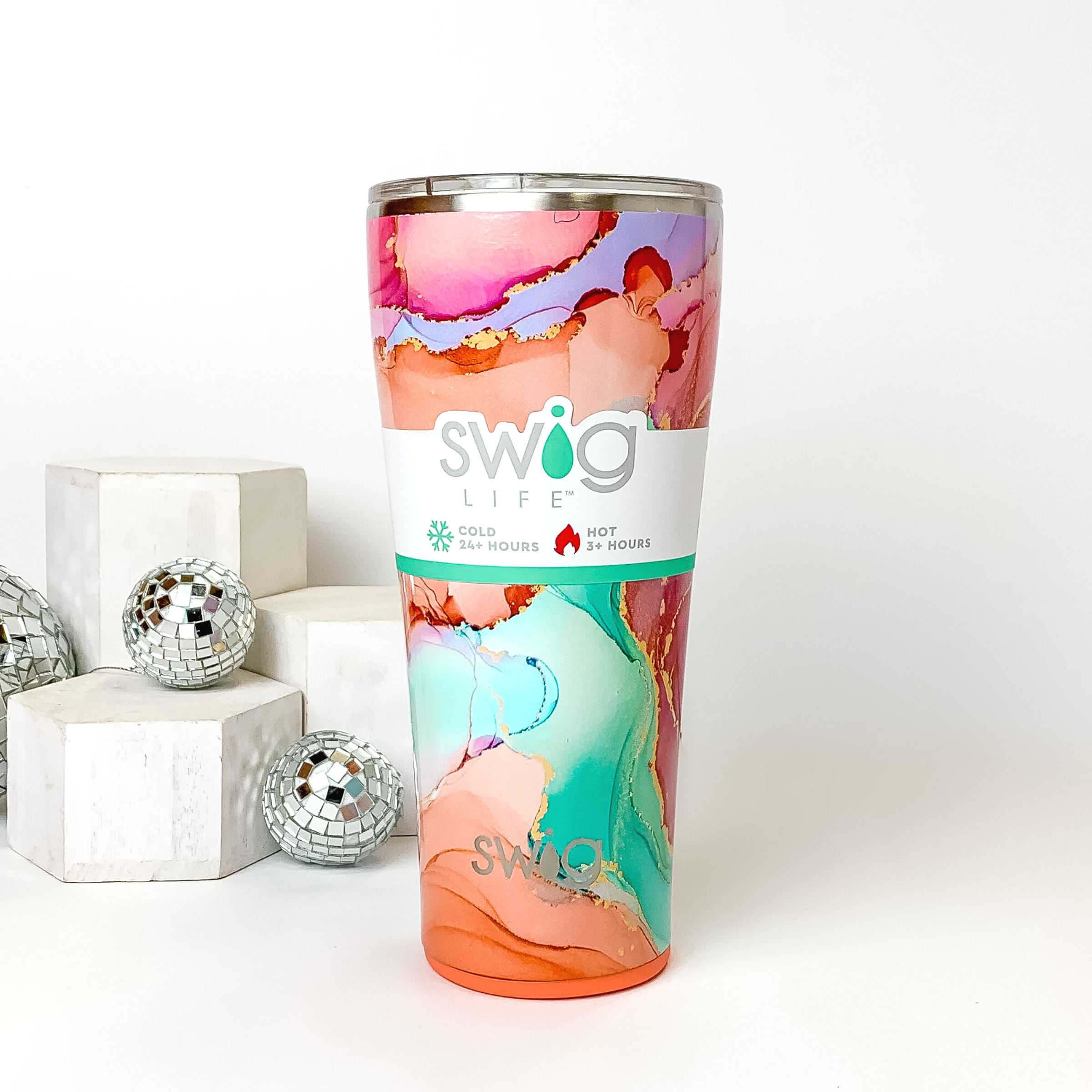 Pink and orange marble tumbler with a clear lid. This marble print includes shades of orange, pink, aqua, and gold glitter. This tumbler is pictured on a white background with white blocks and disco balls on the left side of the picture. 